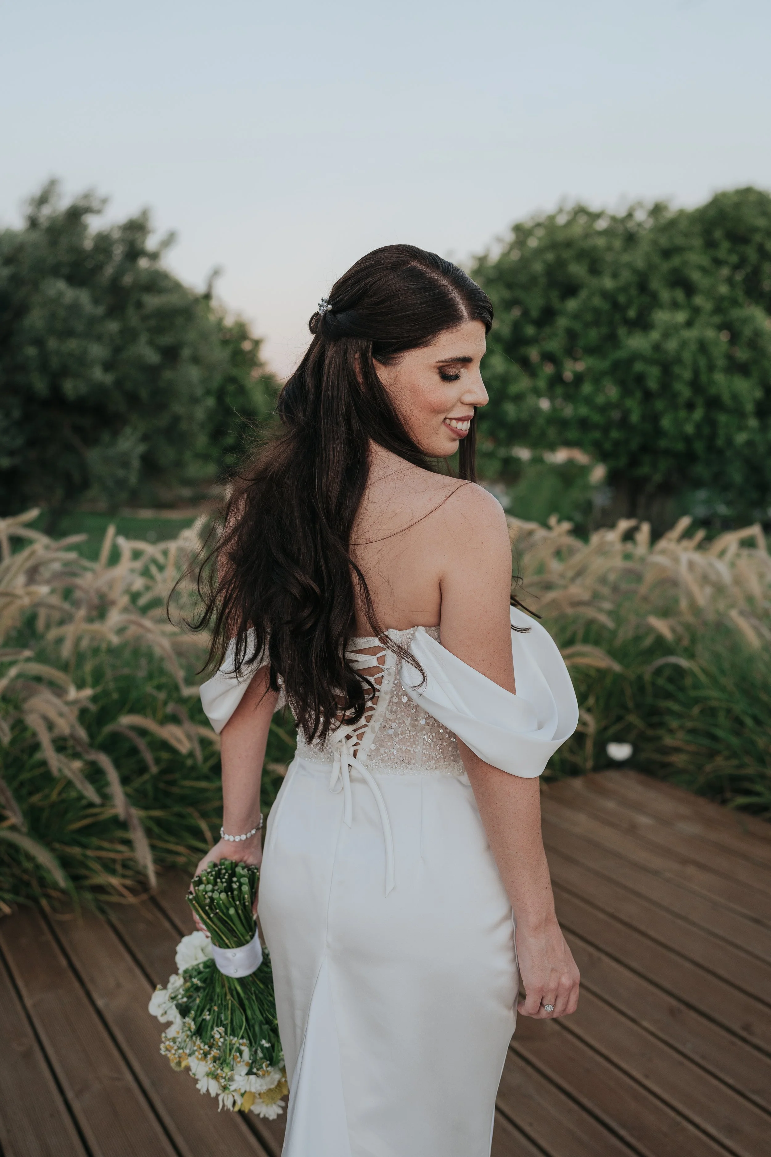 A woman with long, dark hair styled in loose waves, wearing a white gown with off-the-shoulder sleeves and lace-up detail on the side, holds a bouquet of white and green flowers, standing on a wooden deck outdoors with lush green trees and sky in the