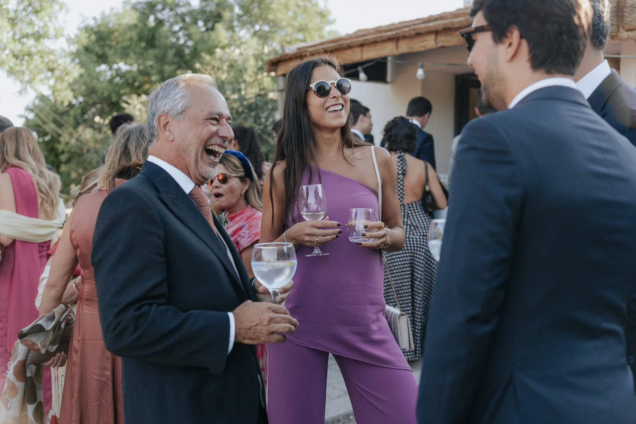 Group of people at a social gathering outdoors, smiling and holding wine glasses, with a woman in purple dress and sunglasses talking to men in suit jackets.