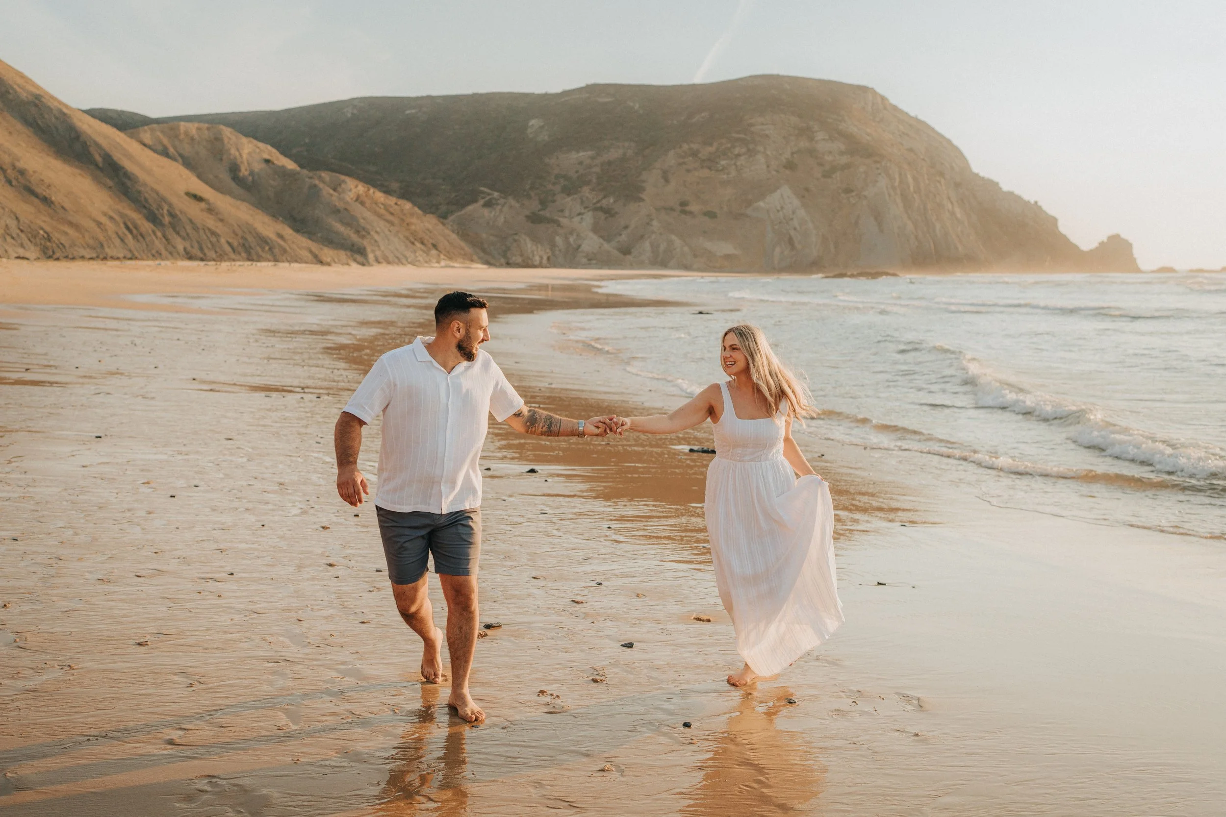 A couple holding hands walking along the sandy beach at sunset, with ocean waves and cliffs in the background.