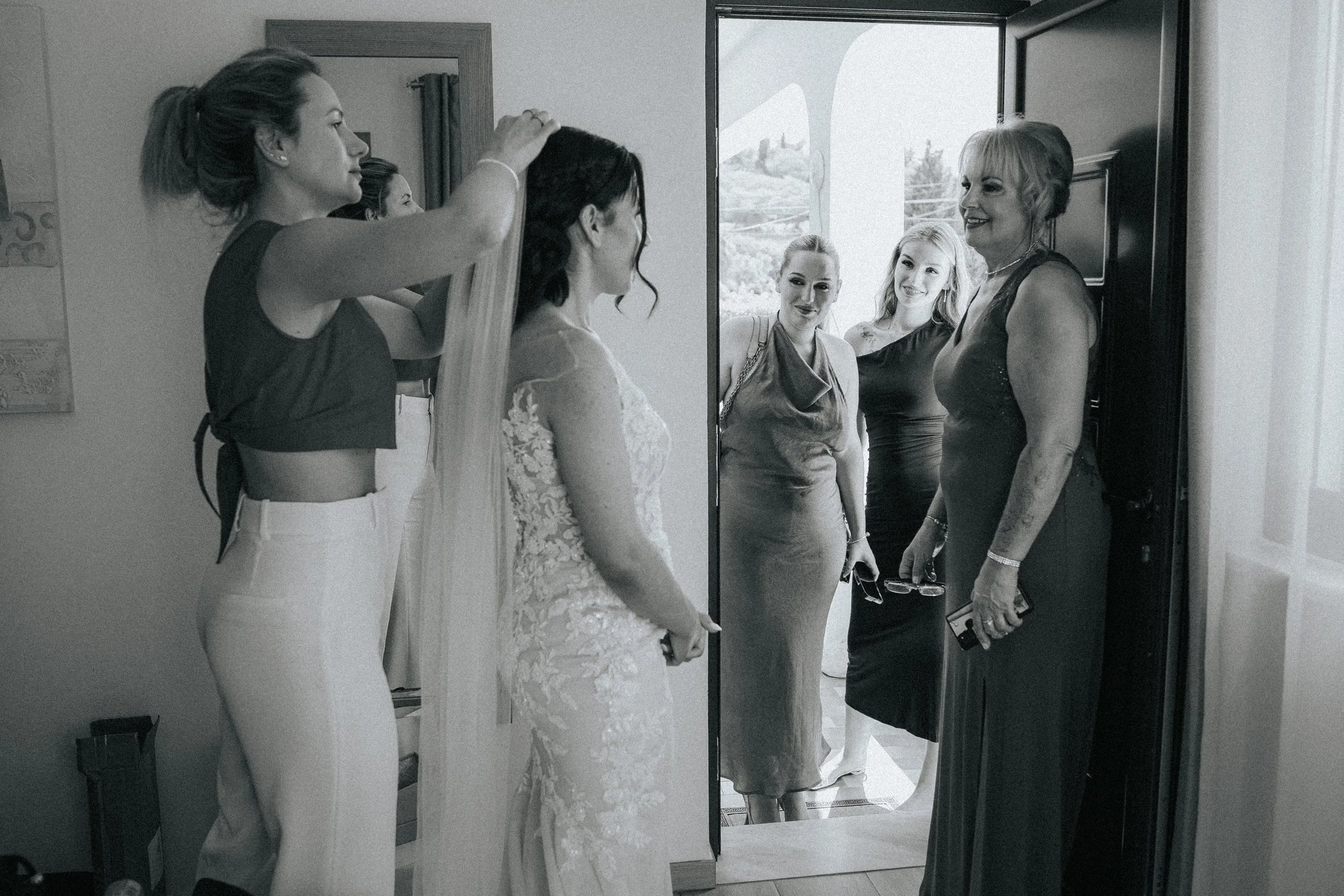 A bride in a lace wedding dress stands in front of a mirror, preparing for her wedding as her bridesmaids assist her. Two women visible in the mirror and one woman outside the mirror are smiling and helping the bride get ready.