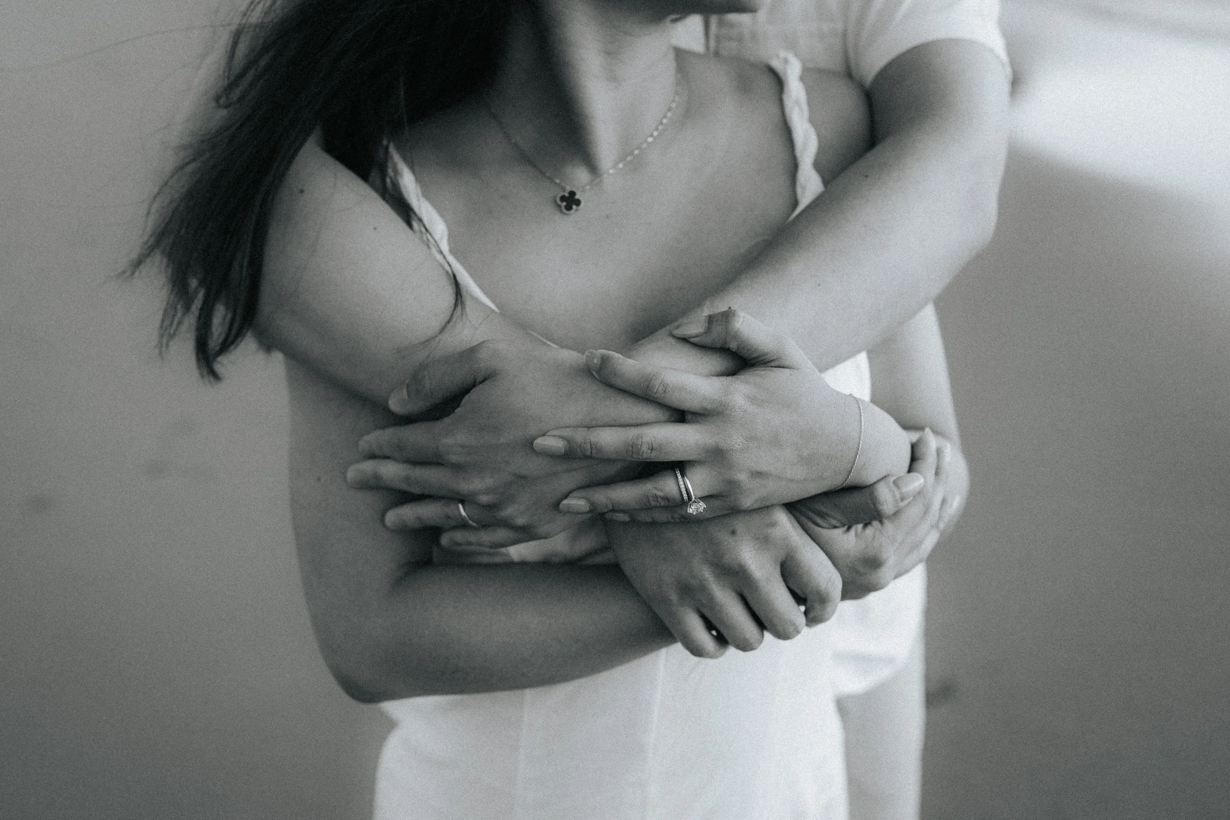 Close-up black and white photo of a woman hugging herself with arms crossed over her chest. She wears a casual white T-shirt, a necklace, and rings on her fingers.