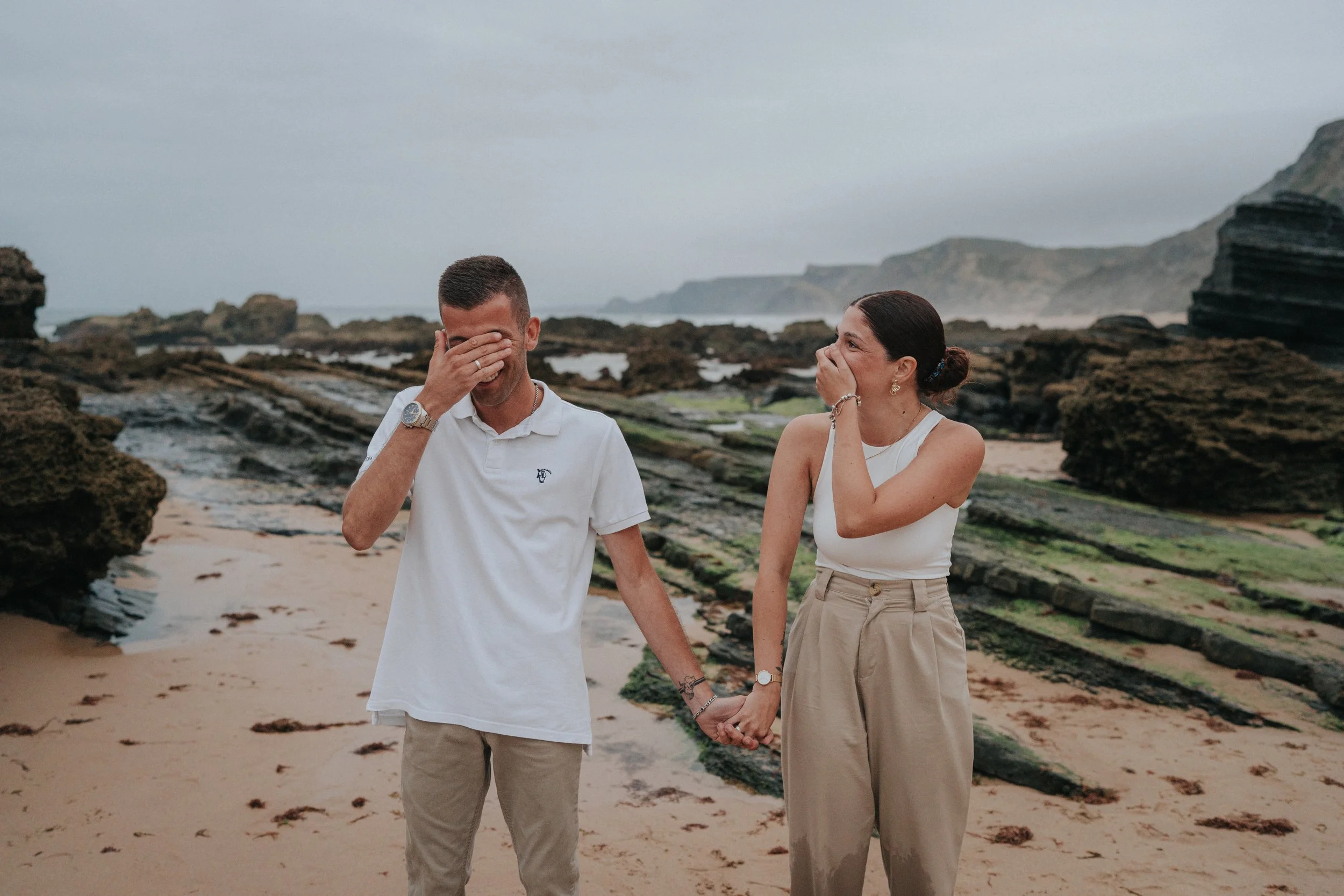 A man and woman stand on a beach with rocky cliffs in the background, holding hands and laughing, with the man covering his face with his hand.