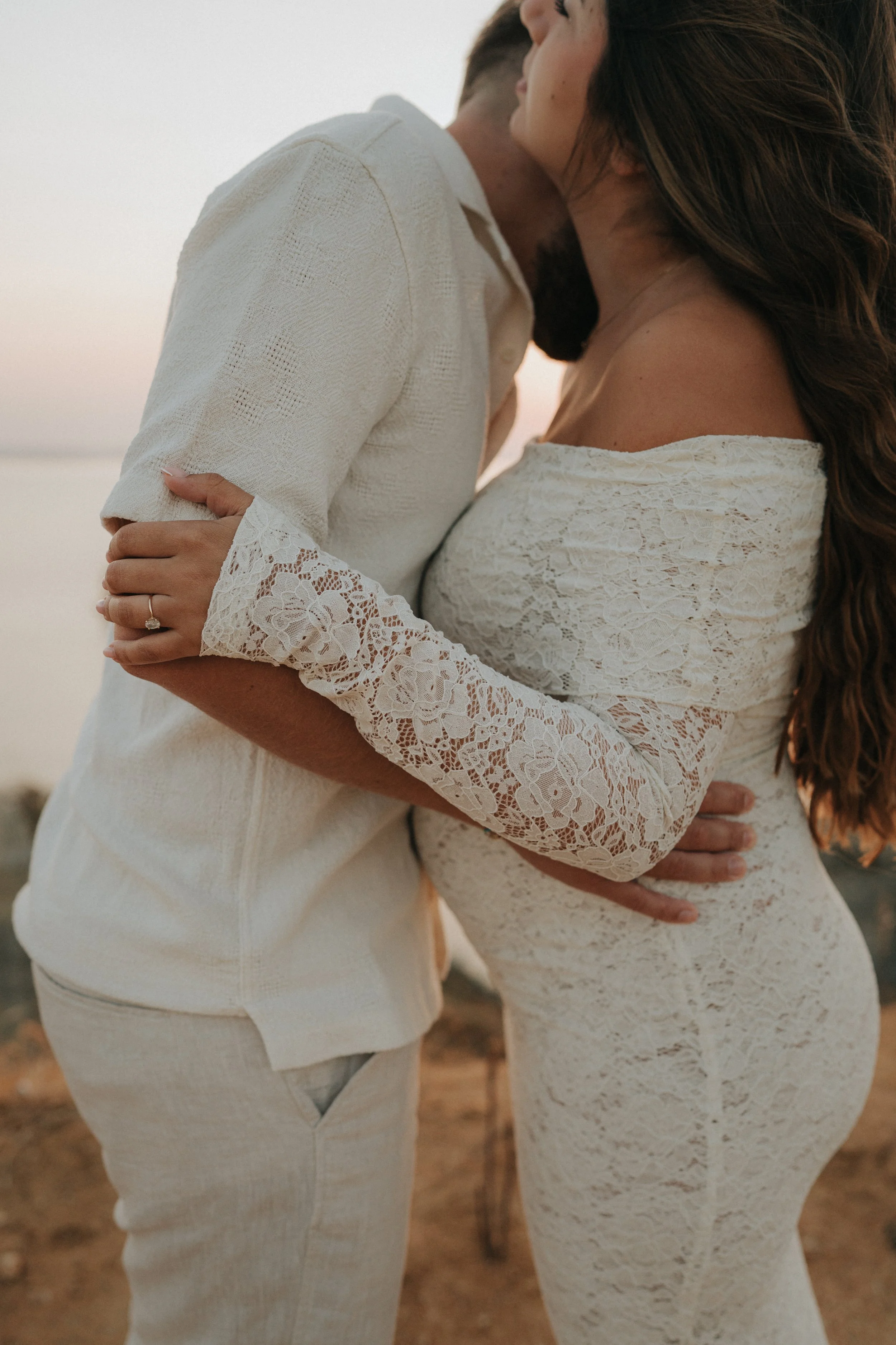 A couple embracing outdoors at sunset, with the woman's hand on the man's chest, both wearing white lace and linen clothing.