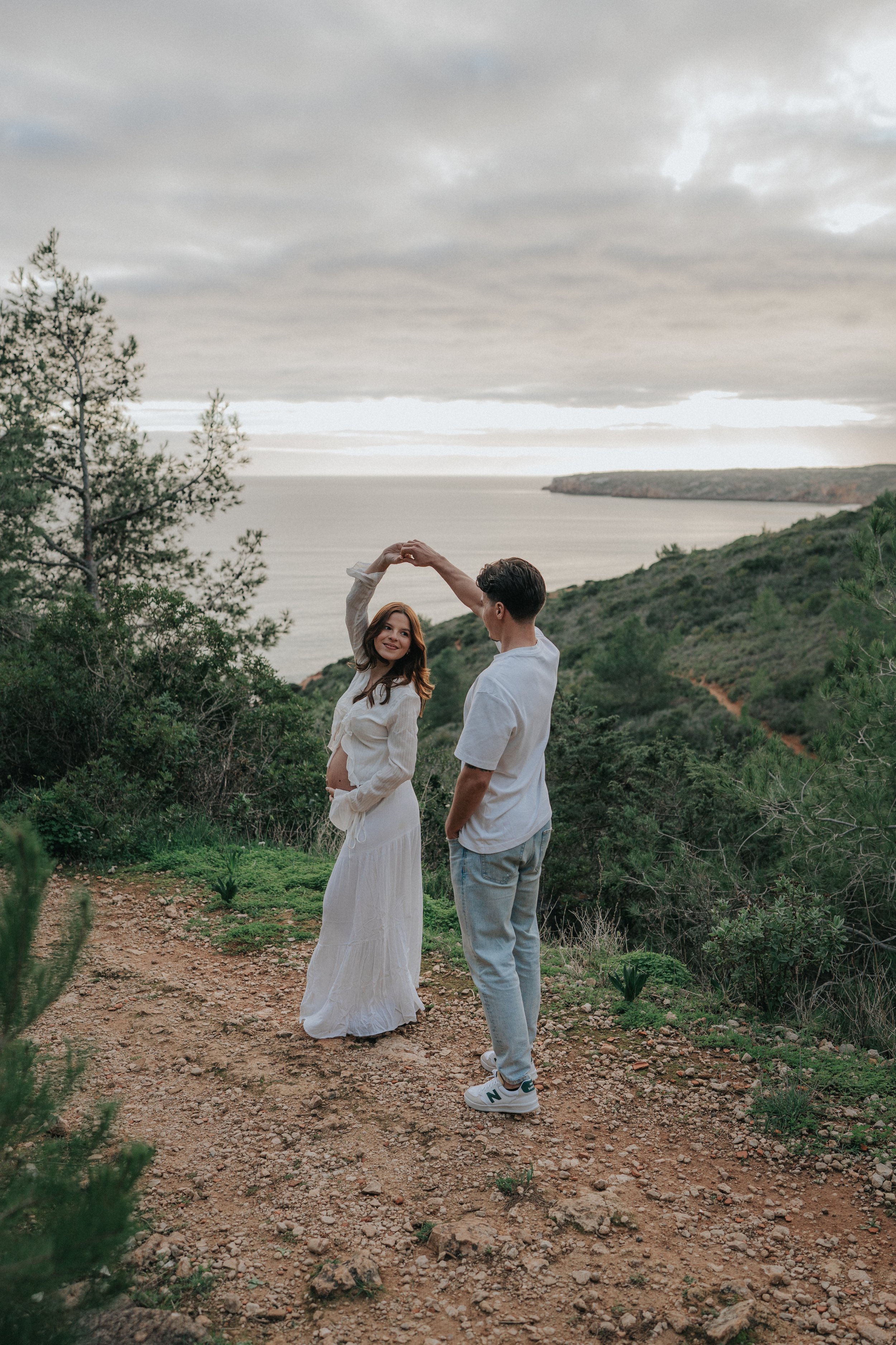 A man and a pregnant woman dancing on a dirt path on a hillside with greenery, trees, and a body of water in the background under overcast sky.