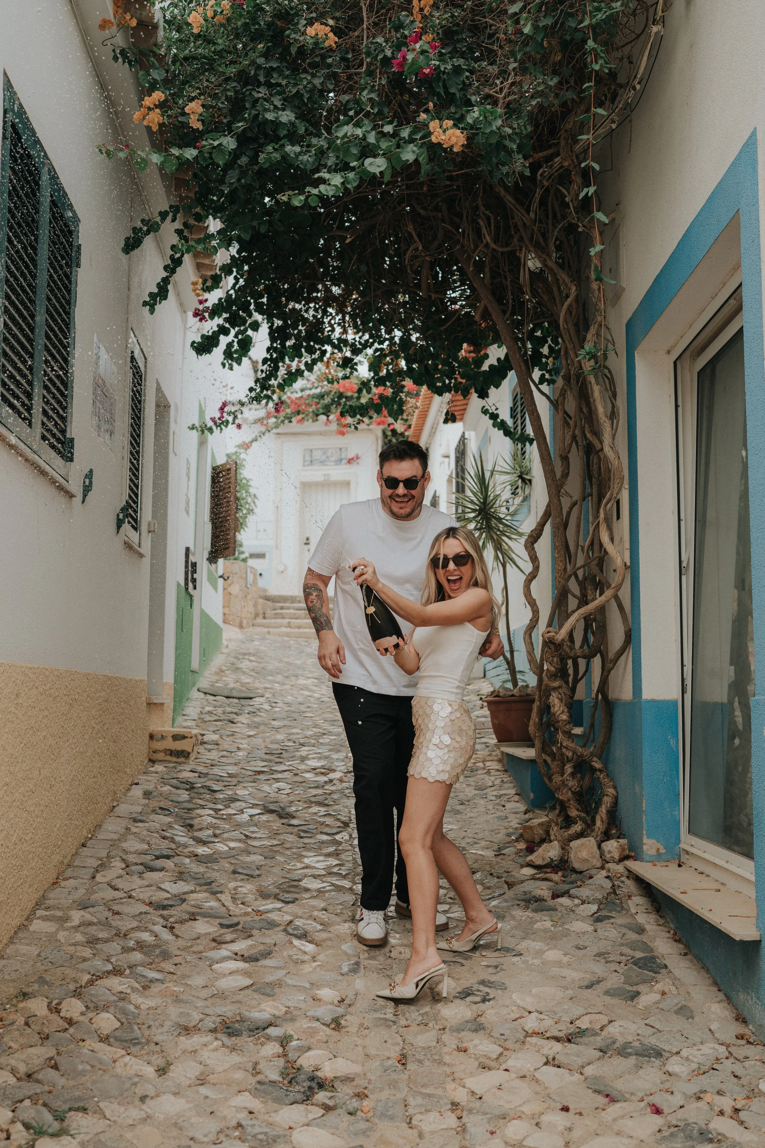 A man and woman celebrating on a cobblestone street, woman holding a champagne bottle, both smiling and wearing casual clothing and sunglasses.