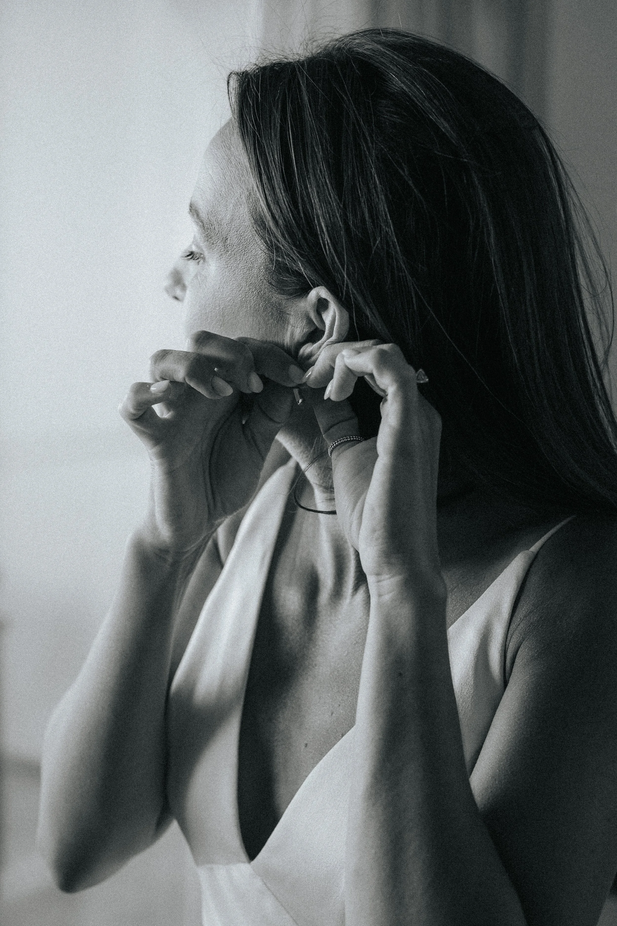 A black and white photo of a woman with long hair, wearing a sleeveless top and earrings, with her hands near her face, appearing contemplative or delicate.