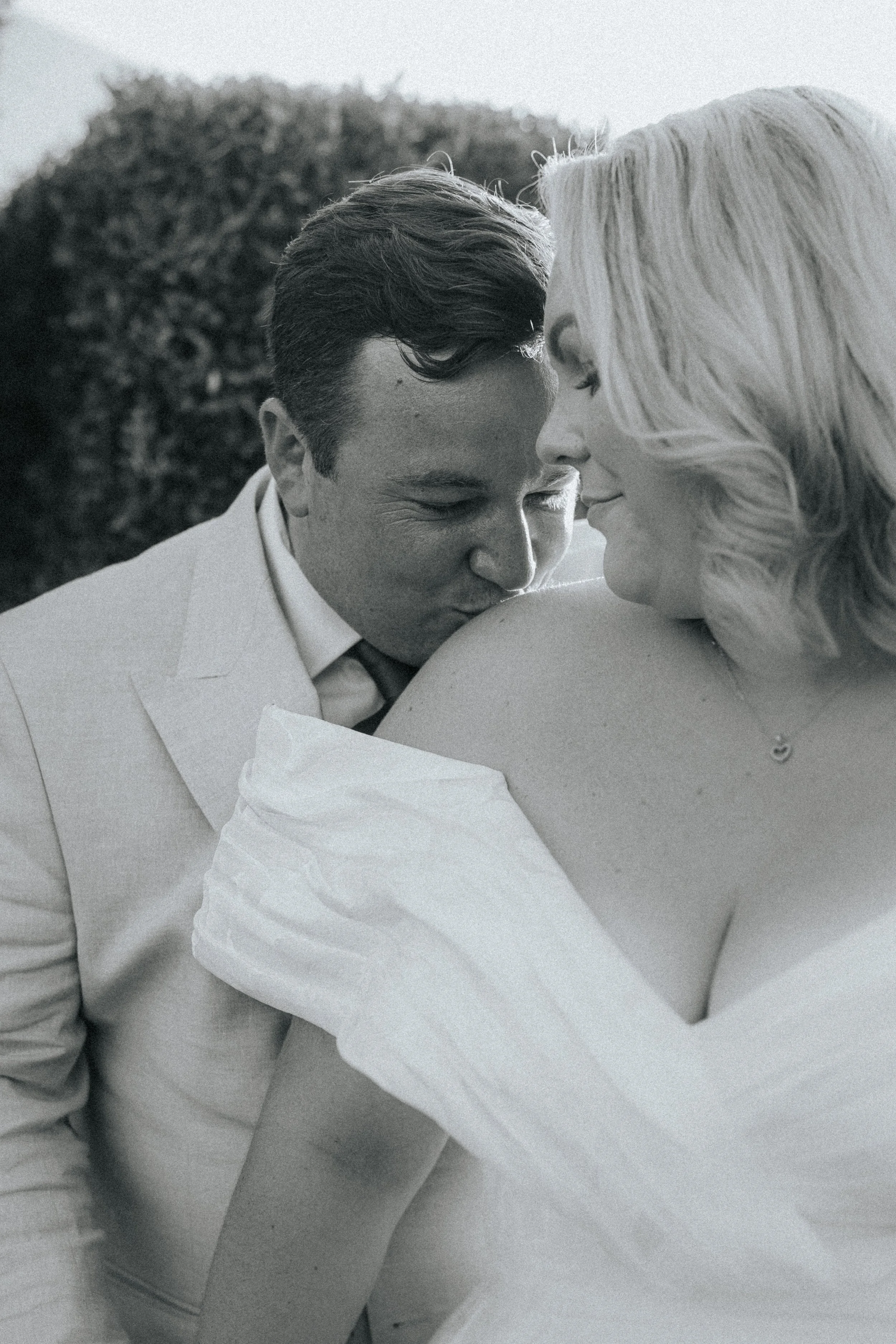 A close-up black and white photo of a man and a woman with their foreheads touching and eyes closed, sharing an intimate moment outdoors with trees in the background.