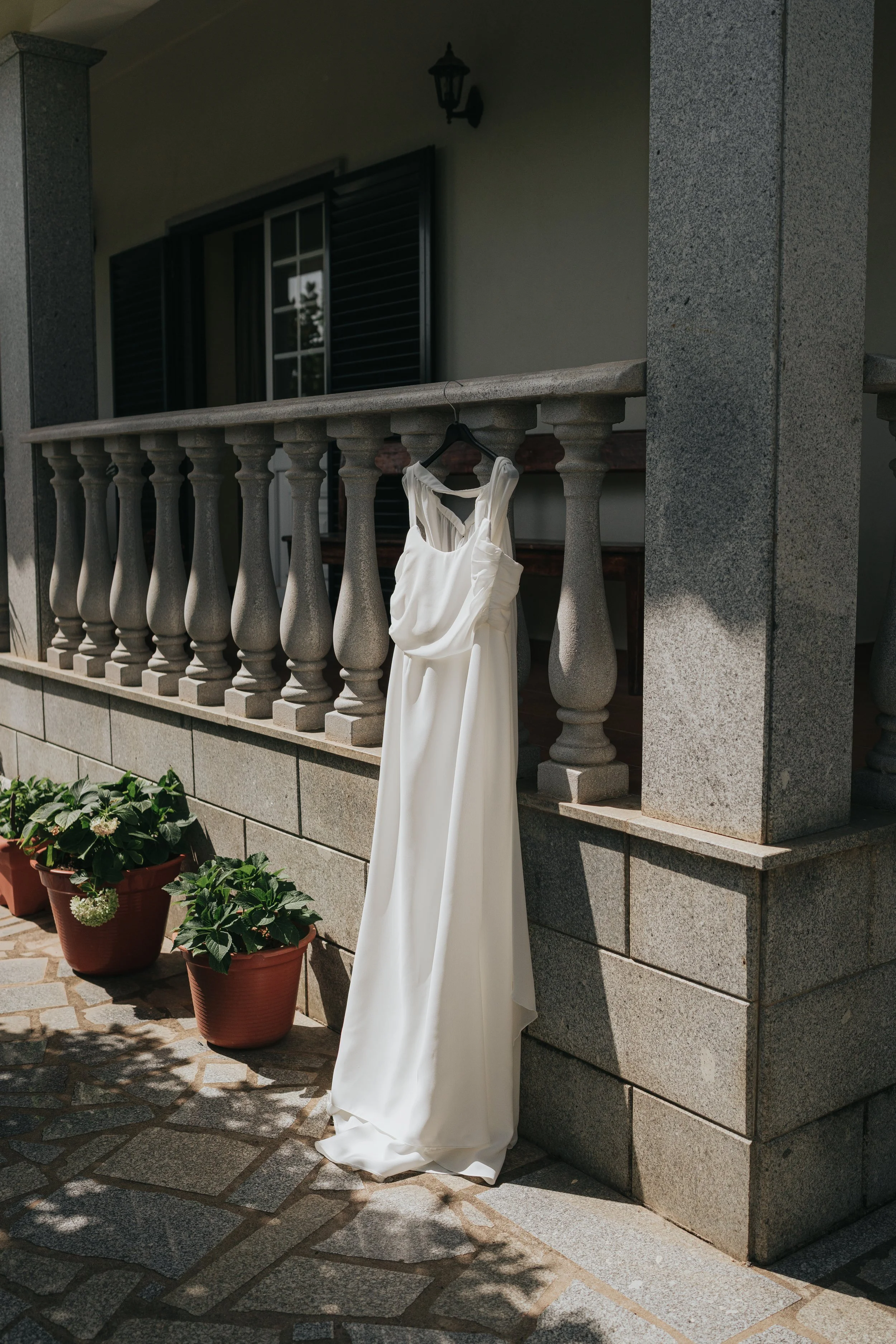 A white dress hangs on a black hanger on a balcony railing, with potted plants below on a stone patio, and a window with black shutters and a lantern above.