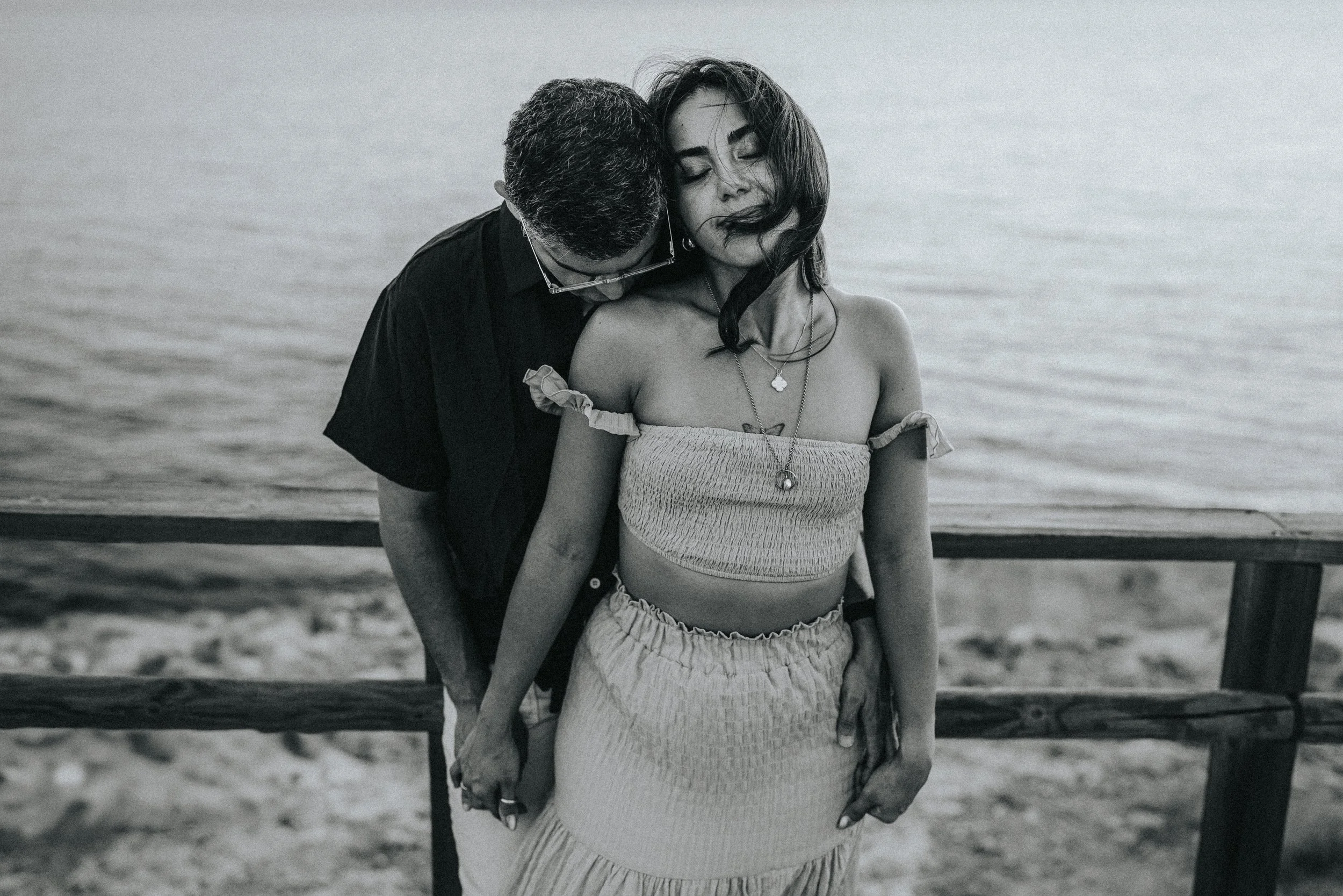 A black and white photo of a man and woman at the beach. The man is leaning his head on the woman's shoulder, and they are close together. The woman has her eyes closed and is wearing a tube top and a skirt. The man has short hair and glasses. They a