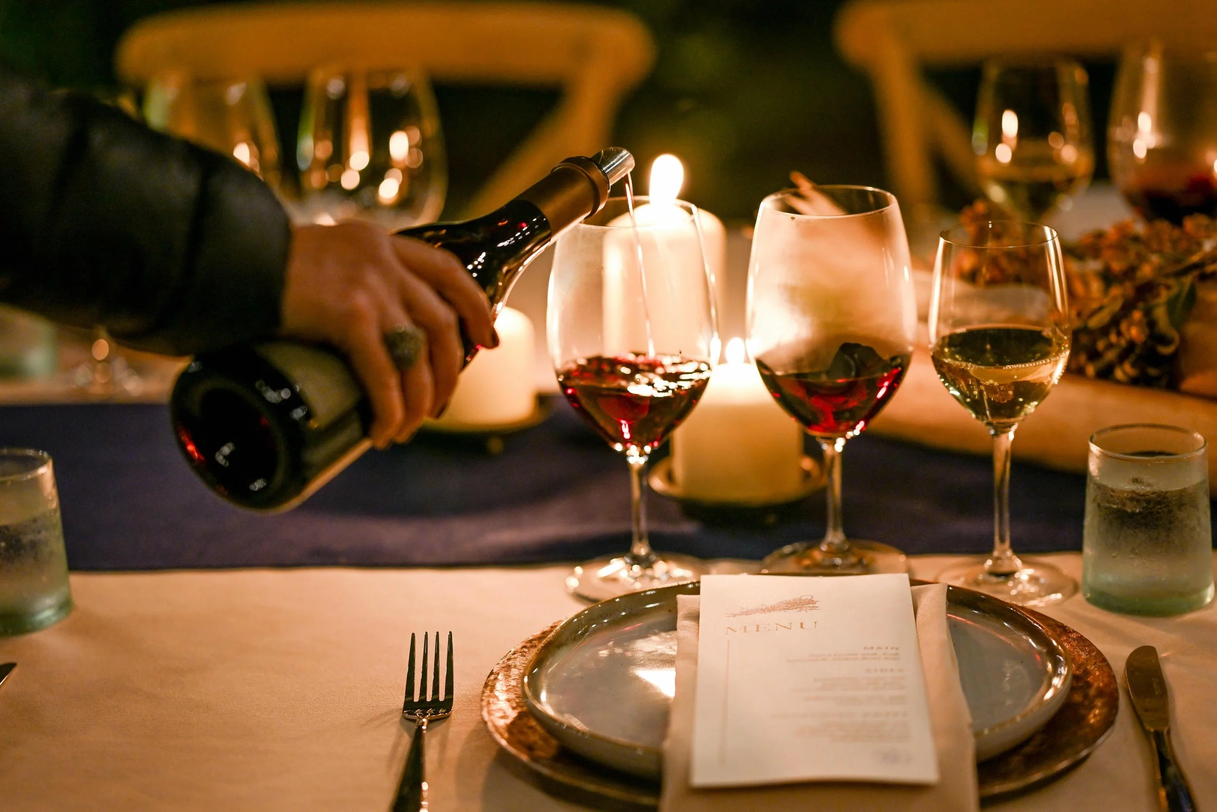 A person pours red wine into a glass at a candlelit dinner table with three glasses of wine, a menu, and a candle.