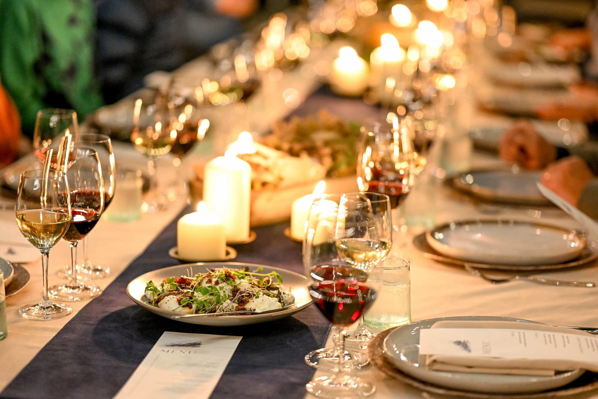A dinner table set with plates, wine glasses filled with white and red wine, surrounded by lit candles and a dark table runner, with a salad in a white bowl as the centerpiece.