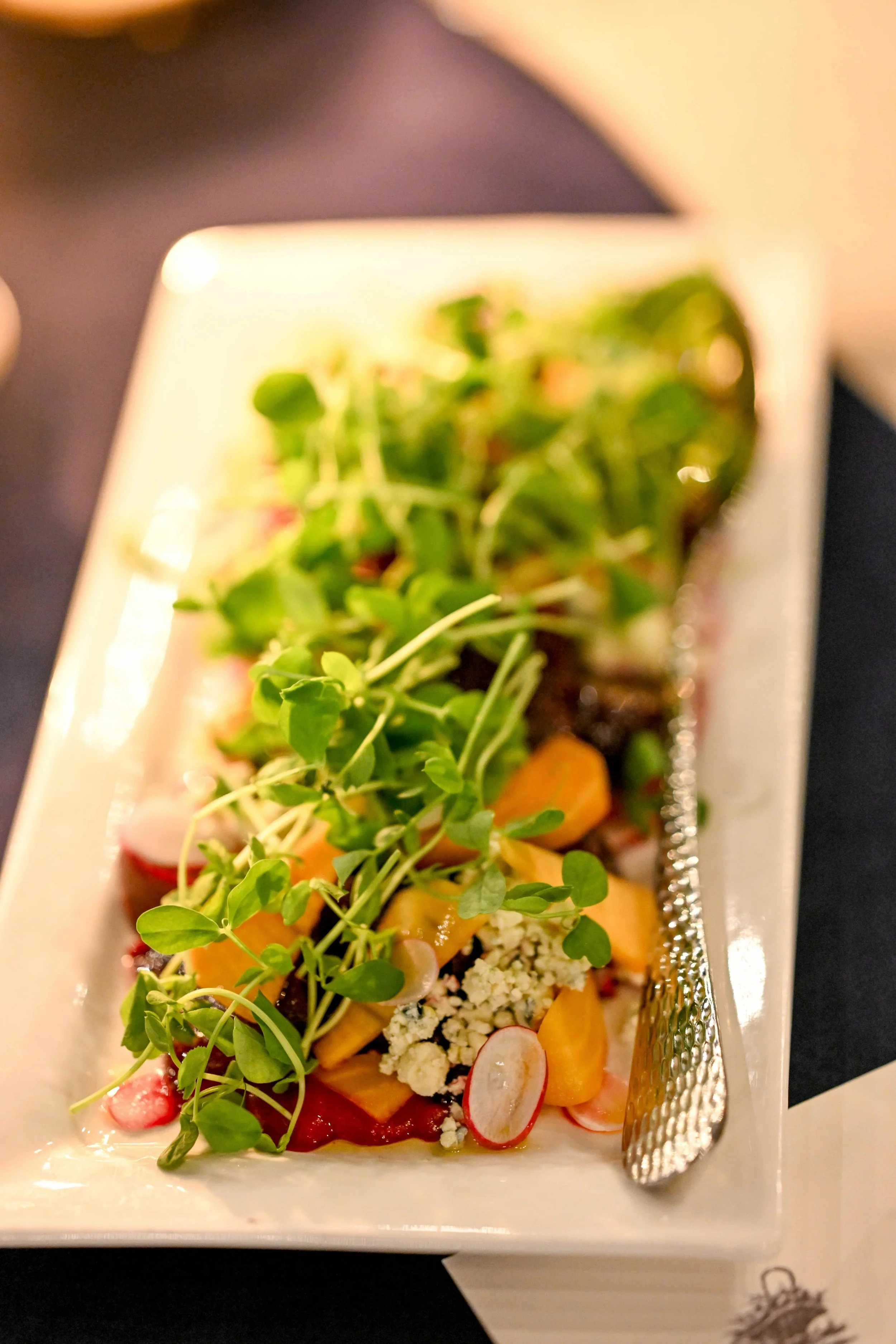 A close-up of a rectangular white plate with a gourmet salad garnished with microgreens, sliced radishes, and crumbled cheese.