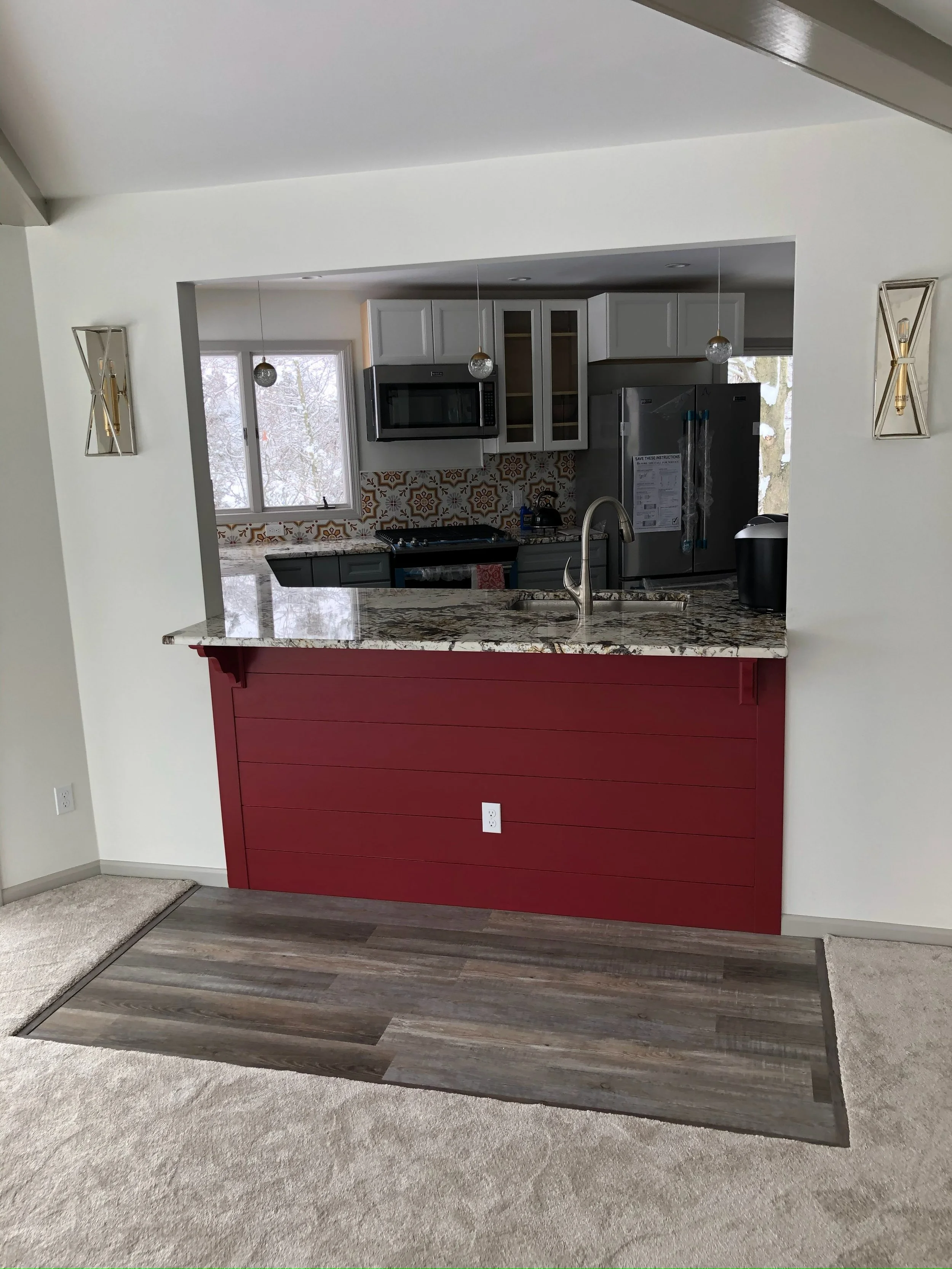 Kitchen with white cabinets, patterned backsplash, and a granite countertop. A pass-through window with a red shiplap bar separates the kitchen from the living area, which has beige carpet and a wooden floor section. Pendant lights hang above the cou