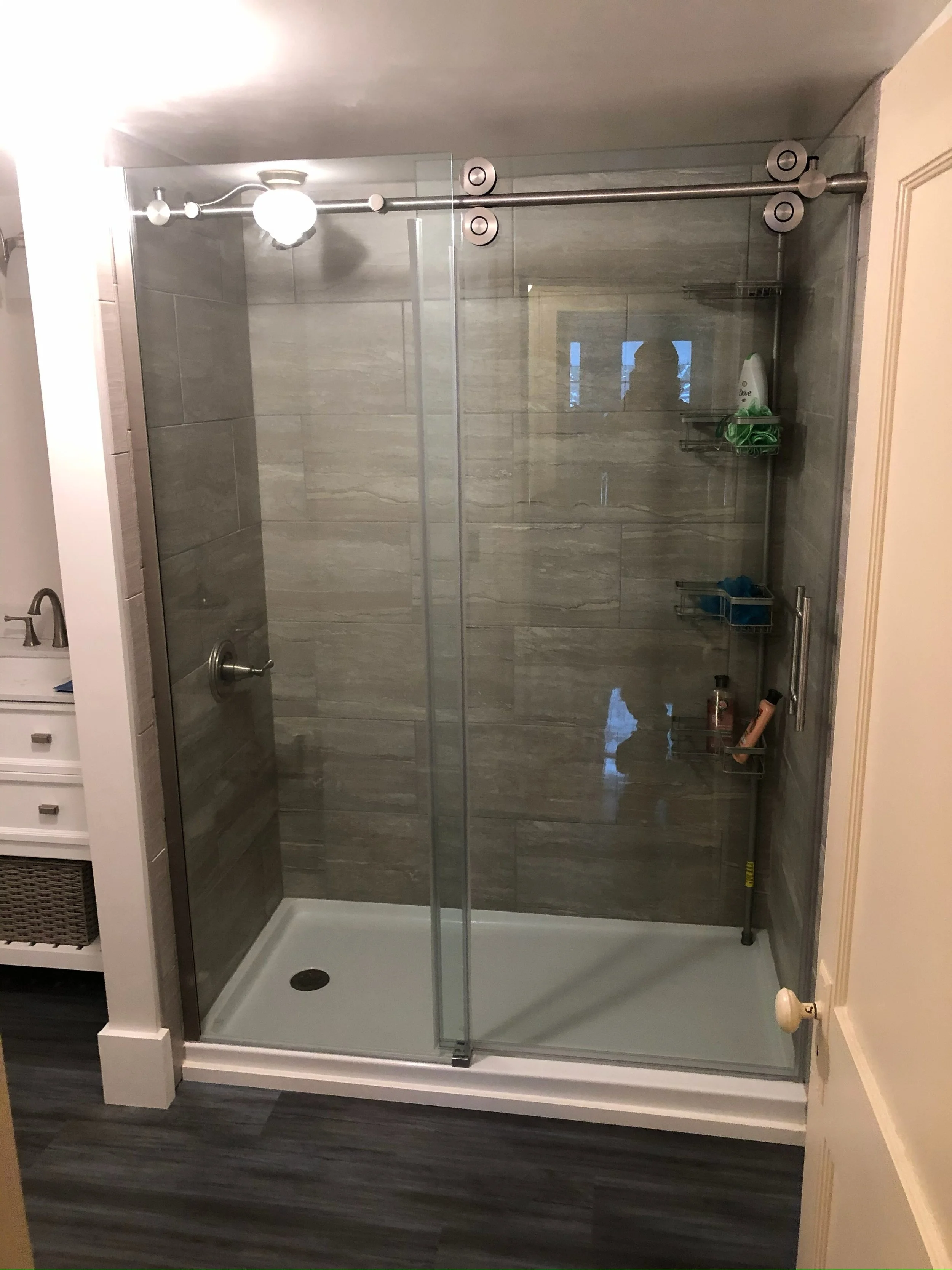 A tiled walk-in shower with glass doors, a handle, and metal shelving for toiletries. The bathroom includes a white cabinet, a sink, and dark flooring.