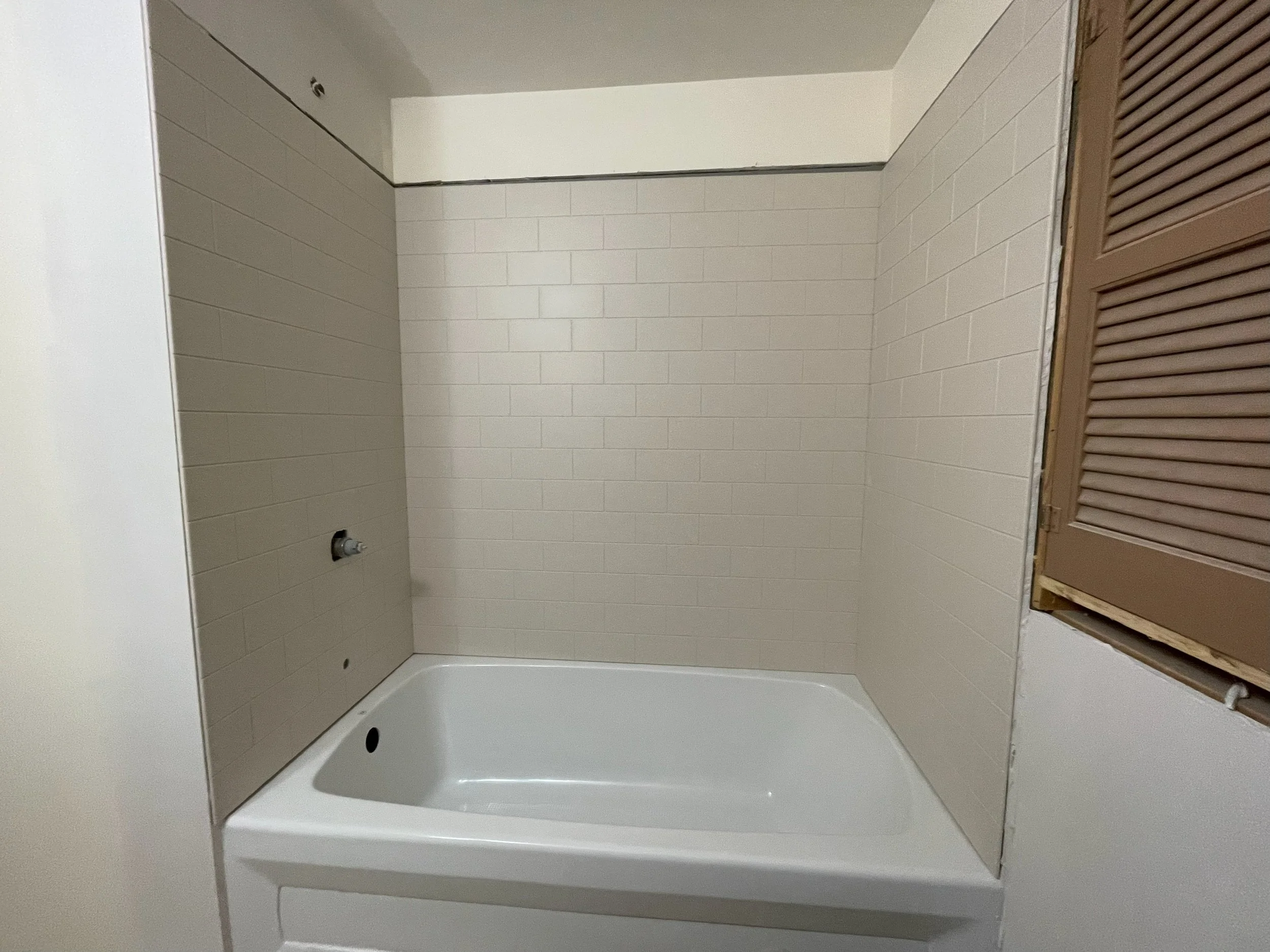 Empty bathtub in a bathroom with beige tiled walls and a window with brown louvered shutters.