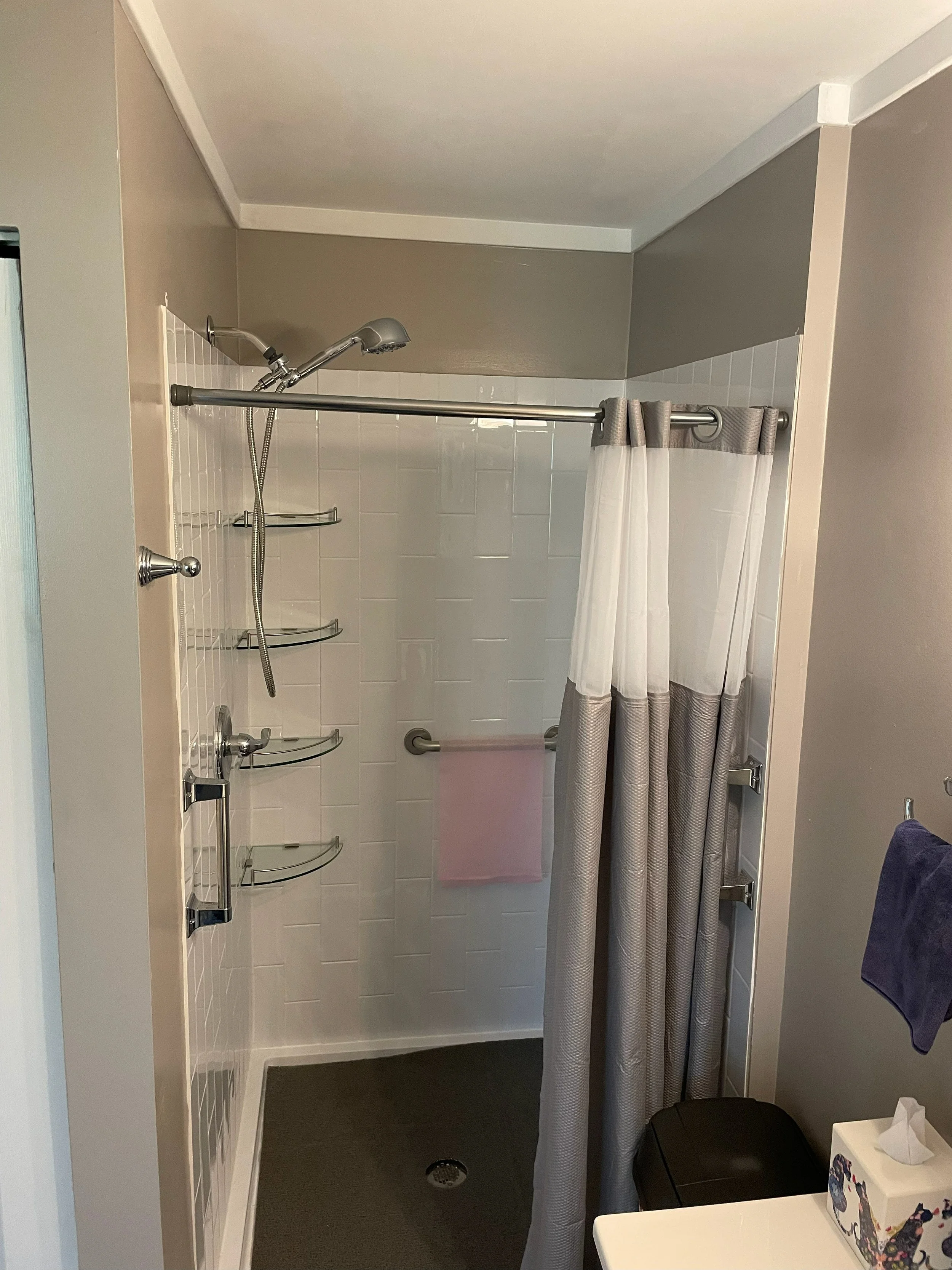 A bathroom shower area with a showerhead, built-in corner shelves, a curtain, a towel rack, and a pink towel. The shower has white tiles and a dark gray floor.