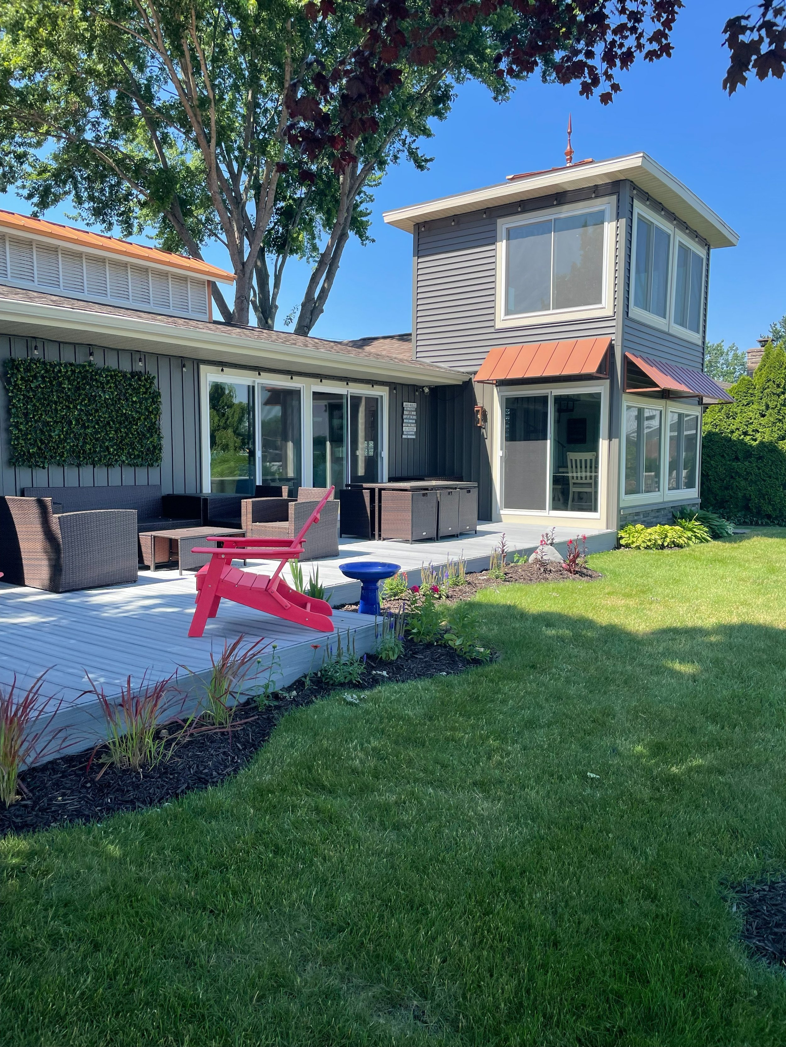 A backyard patio area with outdoor furniture, a red children's rocking chair, and a pink slide, adjacent to a house with gray siding, large windows, and a small garden bed, under a large tree on a sunny day.