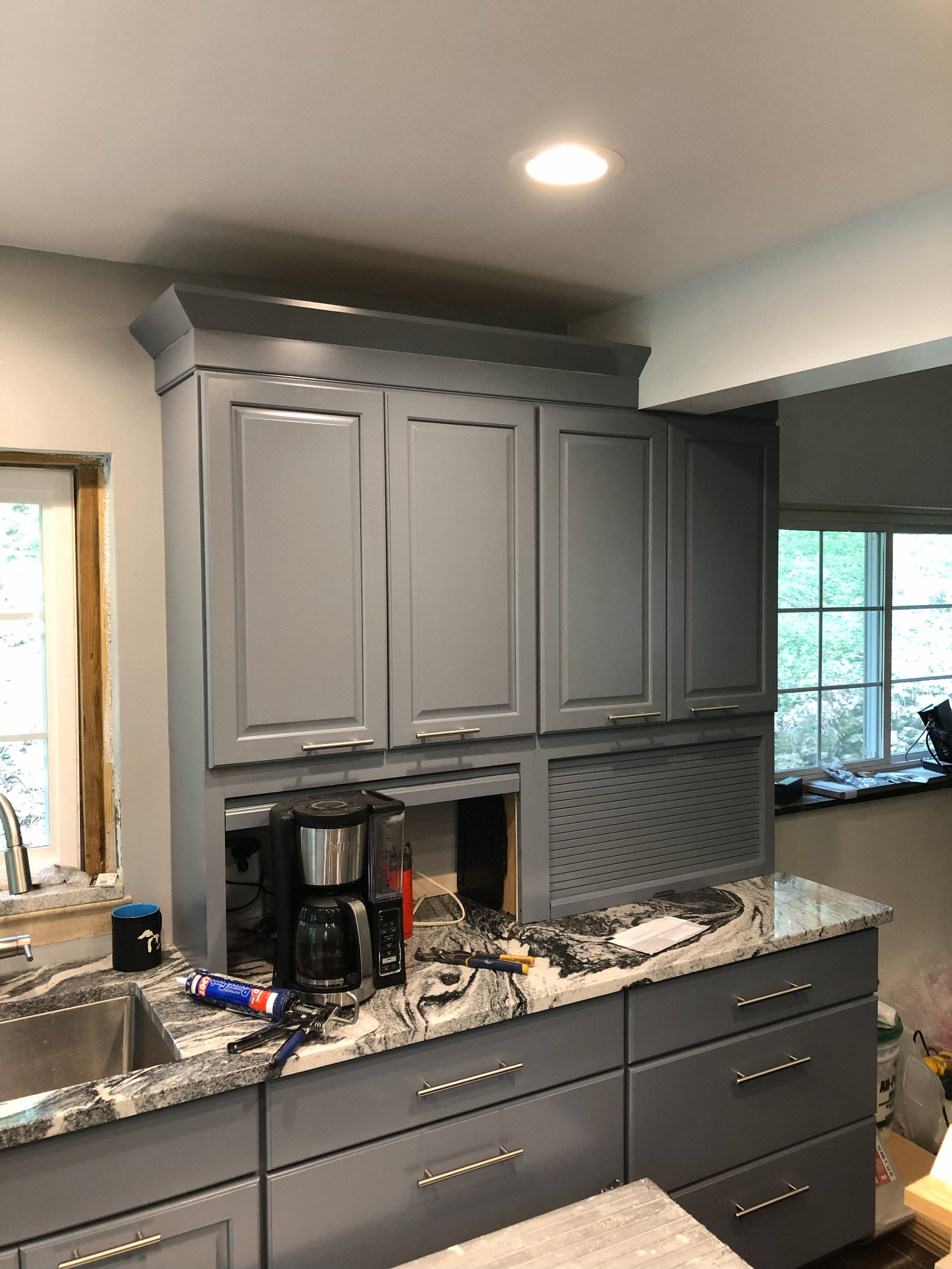 A kitchen with gray cabinets, a marble countertop, and a window. The cabinets are partially installed, with an opening in the wall showing tools and a coffee maker. There is a small black coffee cup on the counter, and tools and materials are scatter