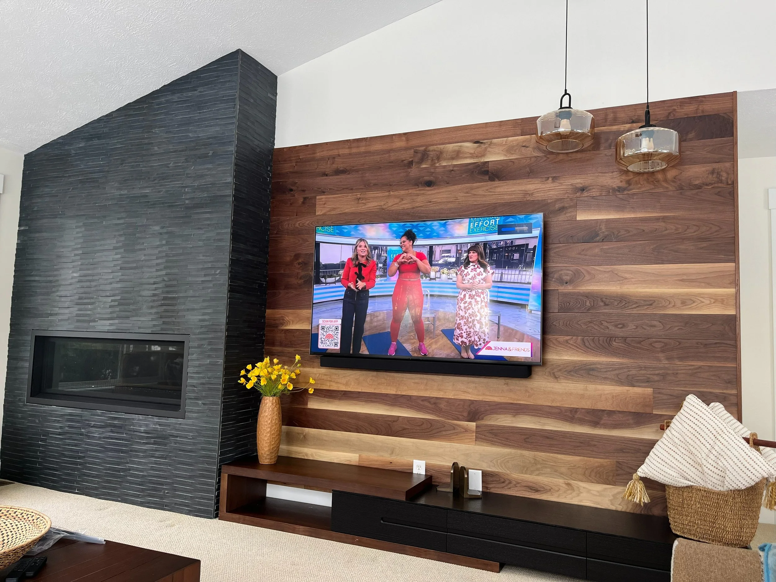 Living room with a black textured fireplace, wooden accent wall, wall-mounted TV showing a talk show with three women, a vase of yellow flowers on a wooden console, and decorative pendant lights hanging from the ceiling.