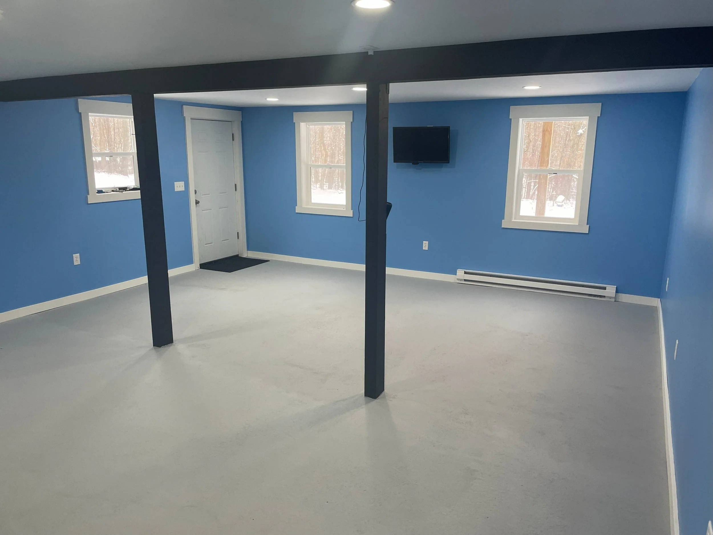Empty room with blue walls, white trim, and three windows showing a snowy outdoor scene; a mounted television on one wall; white carpeted floor; central support beams painted dark blue; white baseboard heater beneath two windows; door with white trim