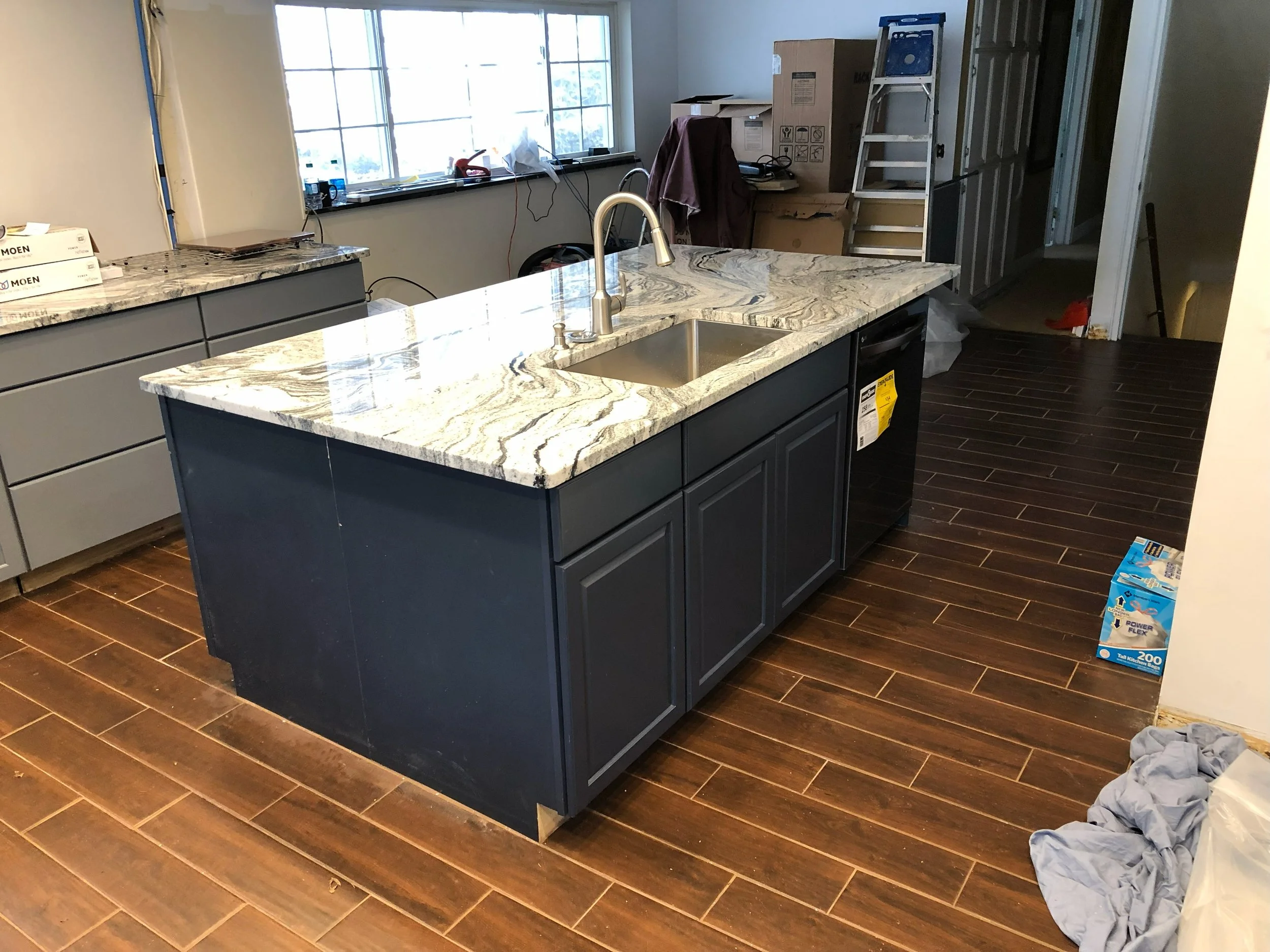 A kitchen with a dark blue island with a marble countertop, a sink, and a dishwasher. There is a window, some boxes, a ladder, and construction supplies in the background.