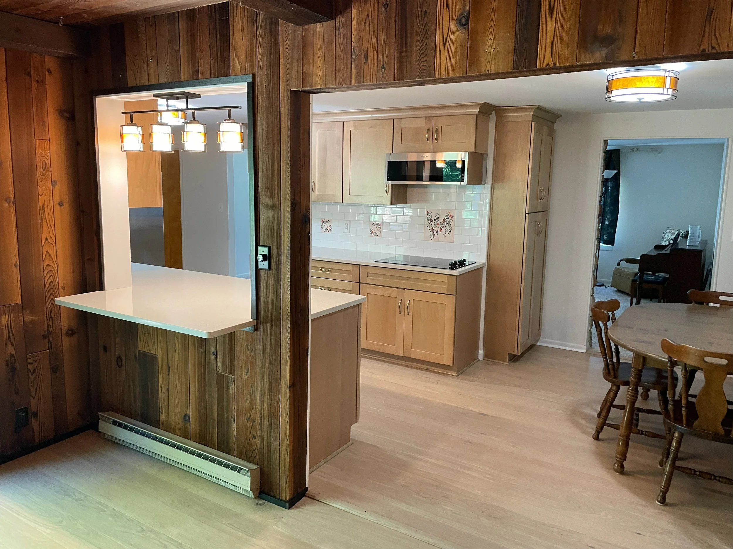 View of a kitchen with wooden cabinets, white tiled backsplash with decorative tiles, a microwave, and a stove. There's a wooden dining table with chairs to the right and a living room with a piano in the background.