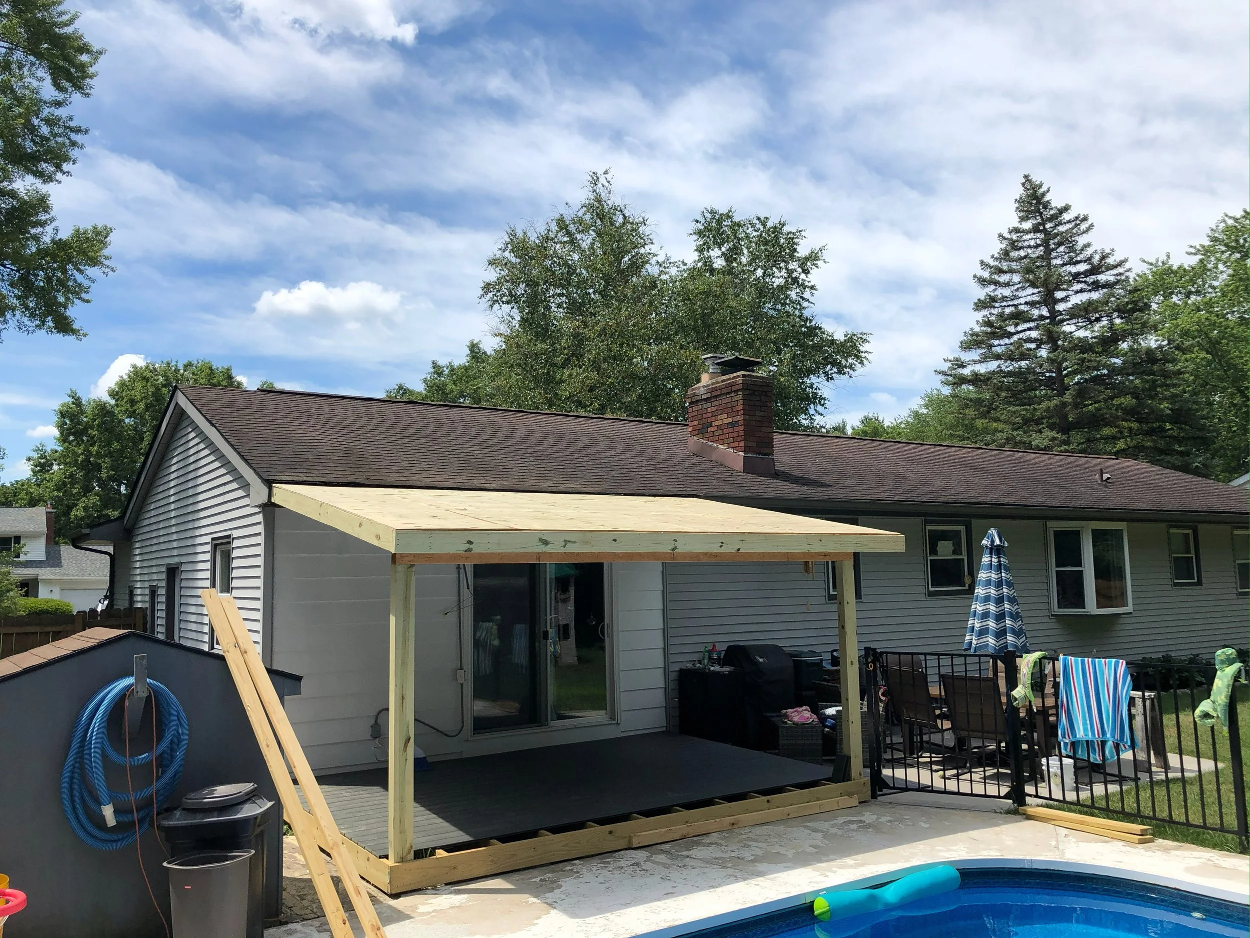Backyard with a house under construction, new wooden deck frame, patio furniture with striped towel, outdoor pool, and trees in the background.