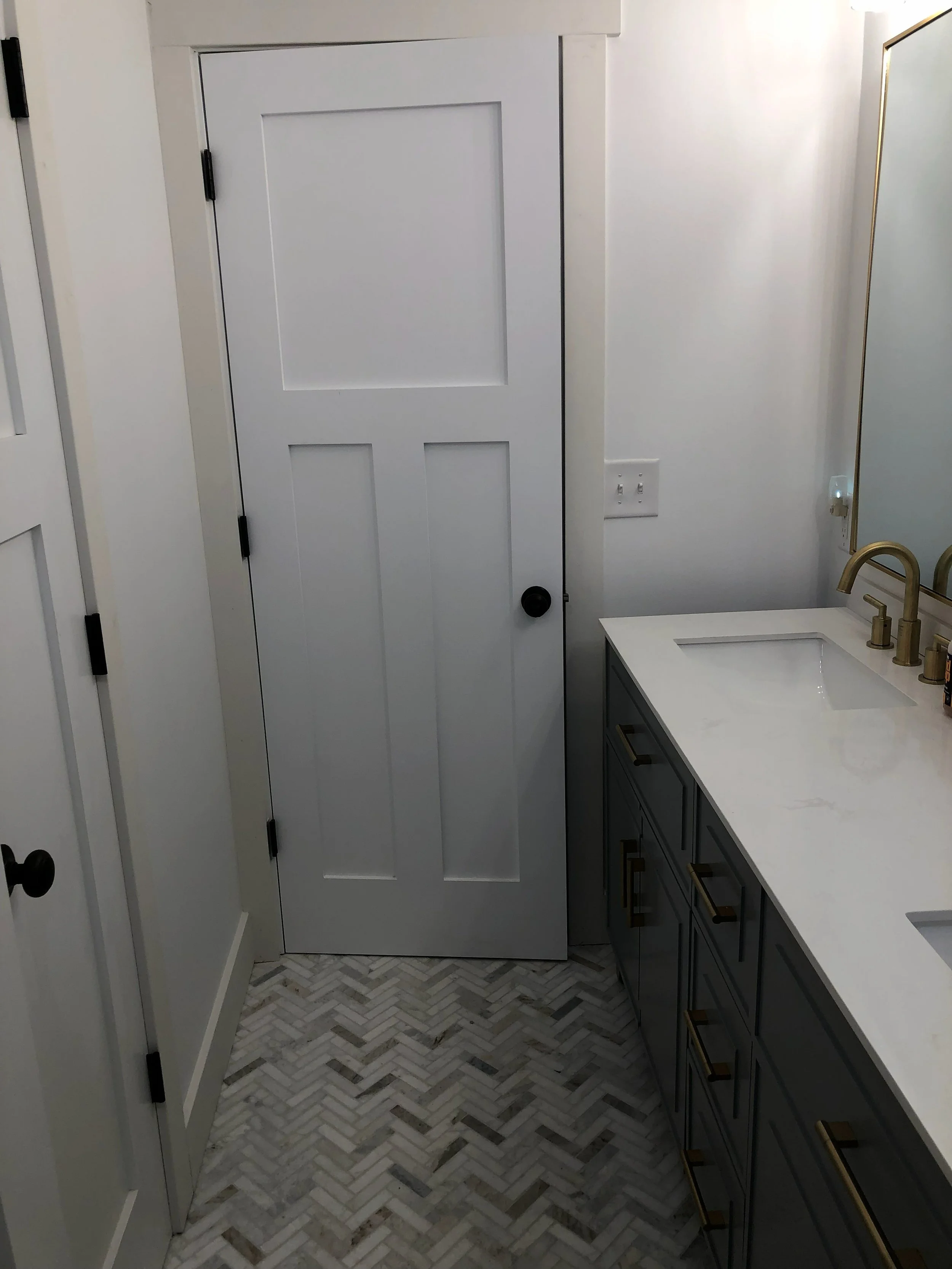 A bathroom with a white wooden door, gray vanity with gold handles, marble countertop with a white sink, and brass faucet. Herringbone patterned gray and white tiles on the floor, white walls, and a large mirror.
