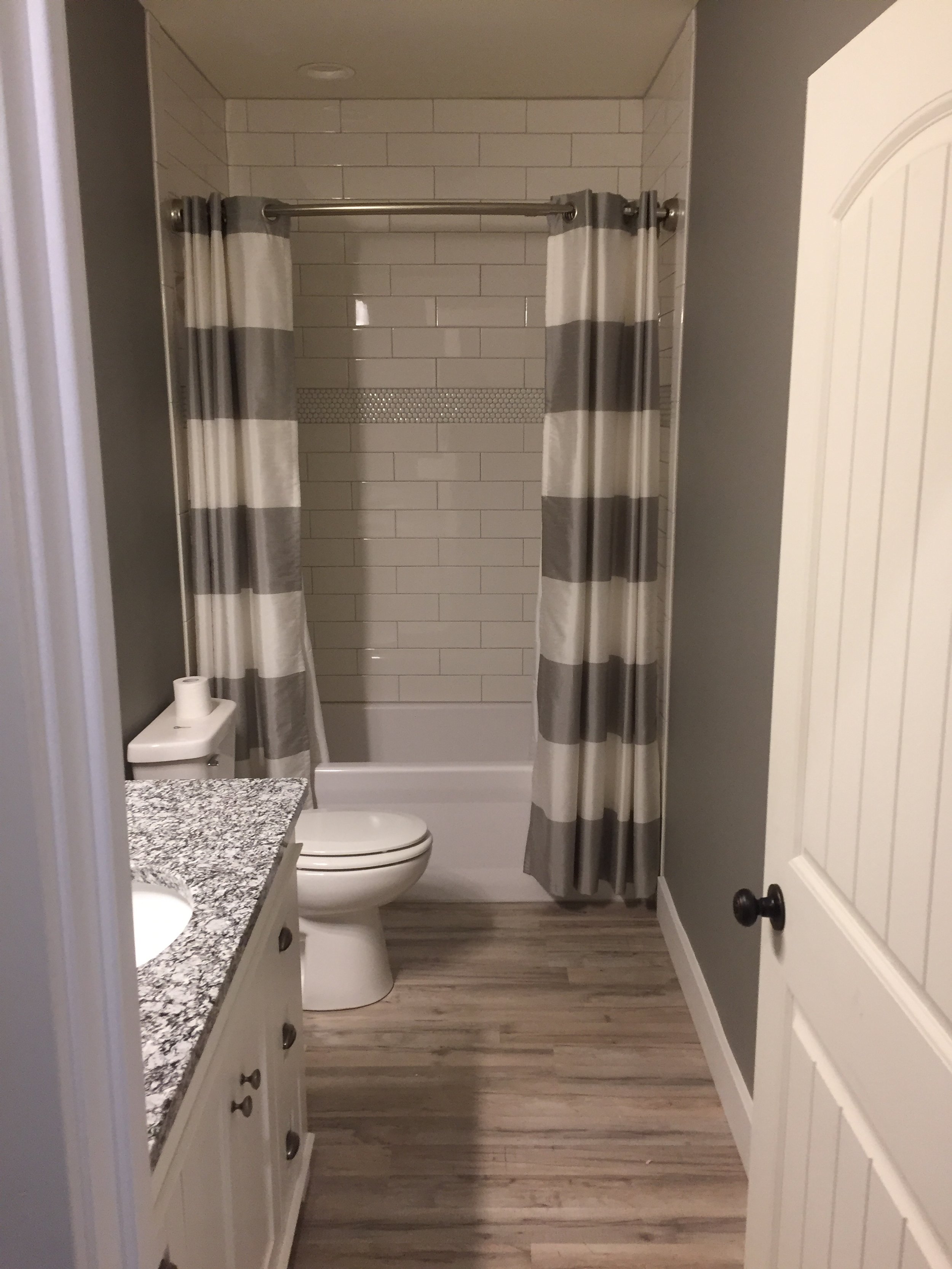A small bathroom with a shower/tub combination, gray and white striped shower curtain, a white toilet, granite countertop with a cabinet, and wood-look vinyl flooring.