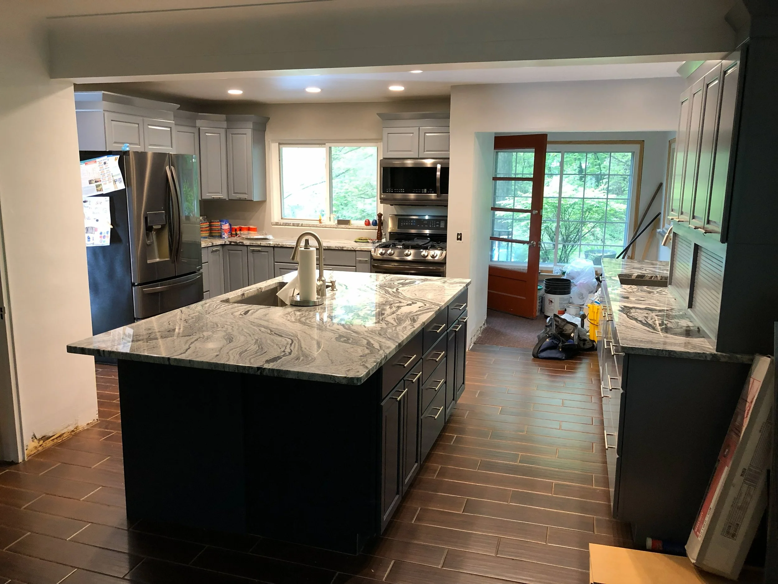 Modern kitchen with gray cabinets, stainless steel appliances, marble countertops, and wooden flooring. An island with a paper towel holder in the center. Large windows and a glass door leading to an outdoor area.
