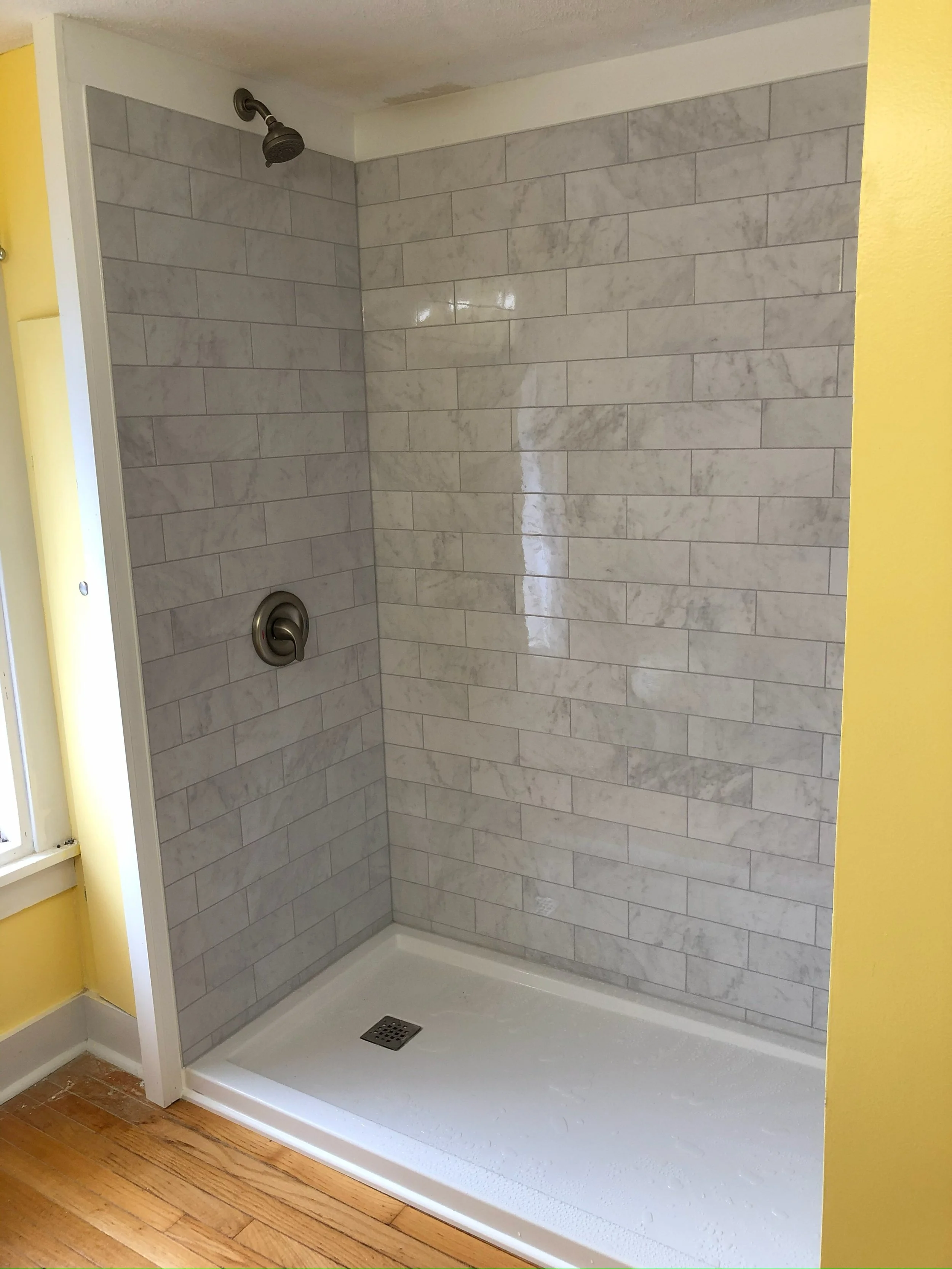Shower stall with gray marble tiles on the walls, a round showerhead, a single handle faucet, and a drain in the white shower base. Part of a yellow wall and a wooden floor are visible.