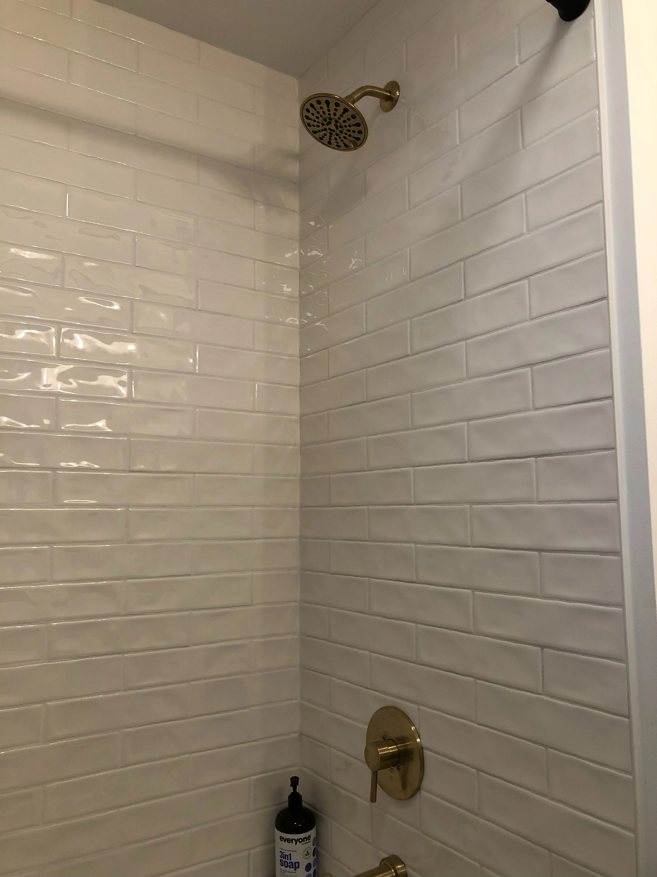Brushed gold showerhead and control valve on white subway tile wall, with a bottle of Everyone 3-in-1 soap on the ledge.