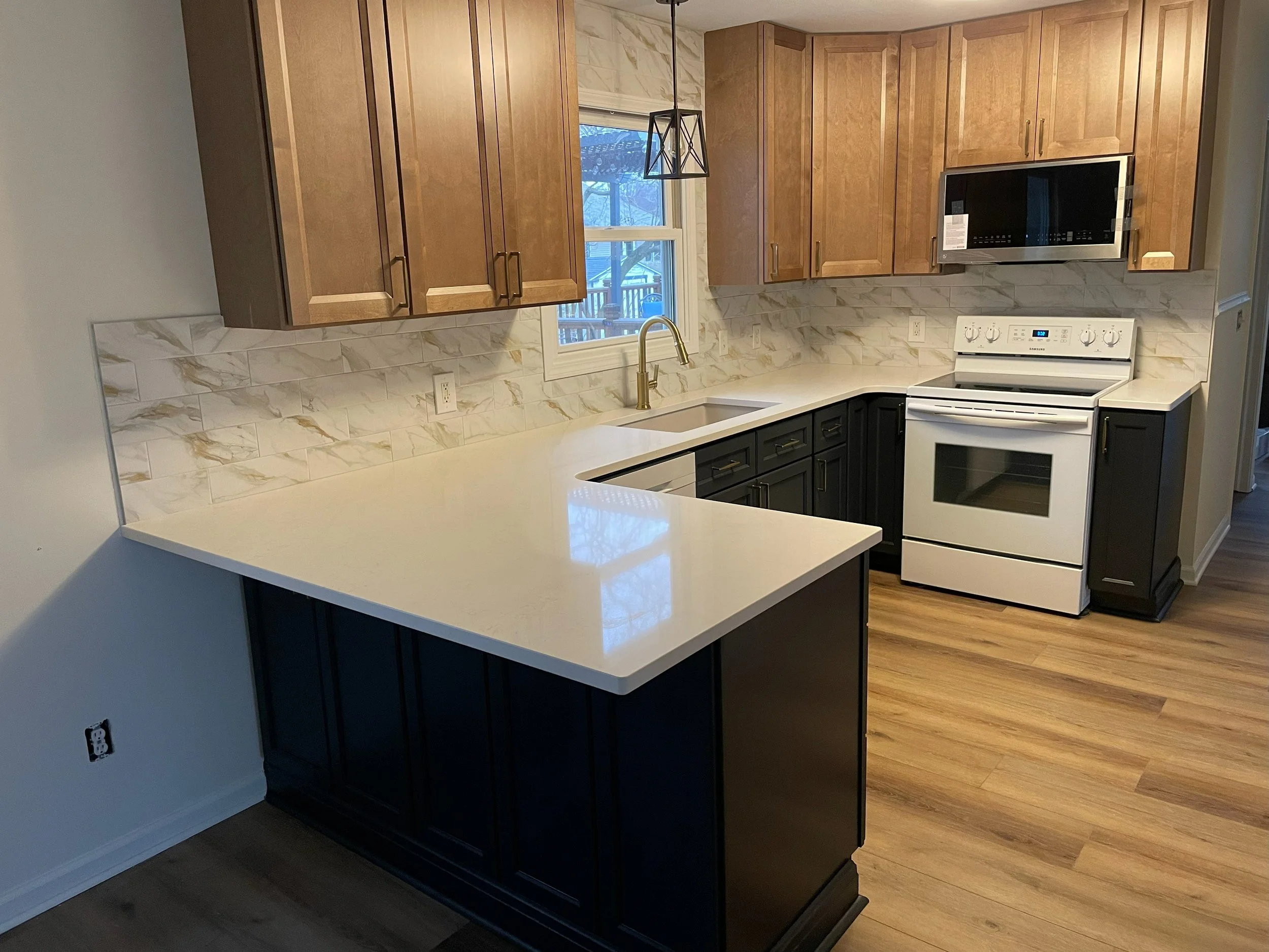 Modern kitchen with wooden upper cabinets and black lower cabinets, white countertops, a window above the sink, marble-style backsplash, and white stove with microwave above.