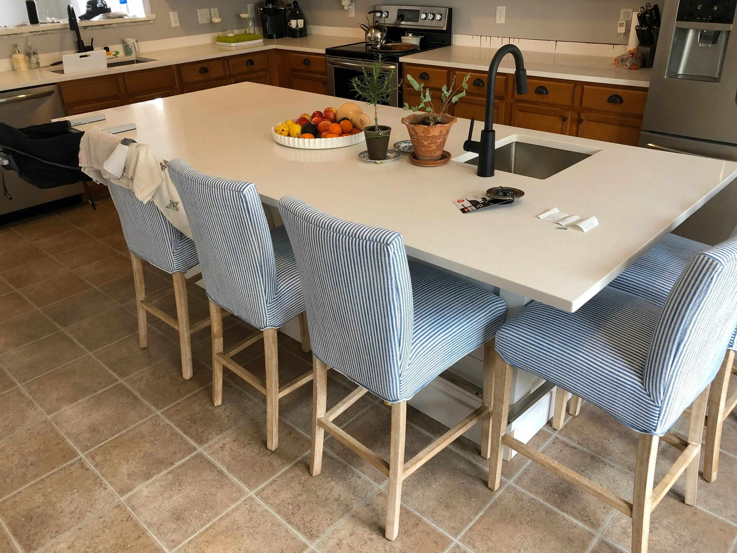 A kitchen with a large central island countertop surrounded by six blue and white striped chairs. On the countertop, there are potted plants, a fruit bowl with various fruits, and a sink with a black faucet. The background features wooden cabinets, a