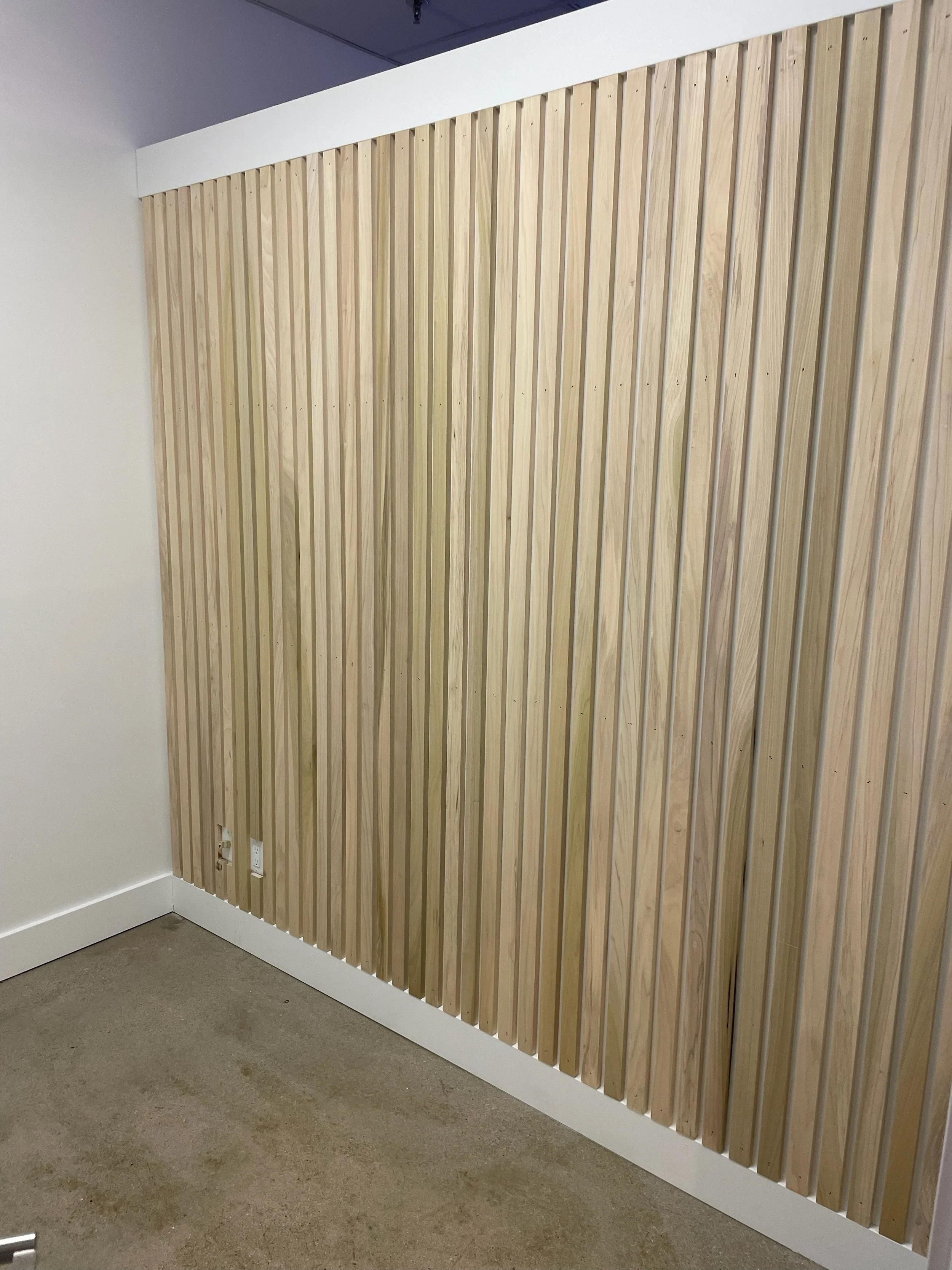 Interior wall with vertical wooden slats, white baseboard and electrical outlet near the floor.