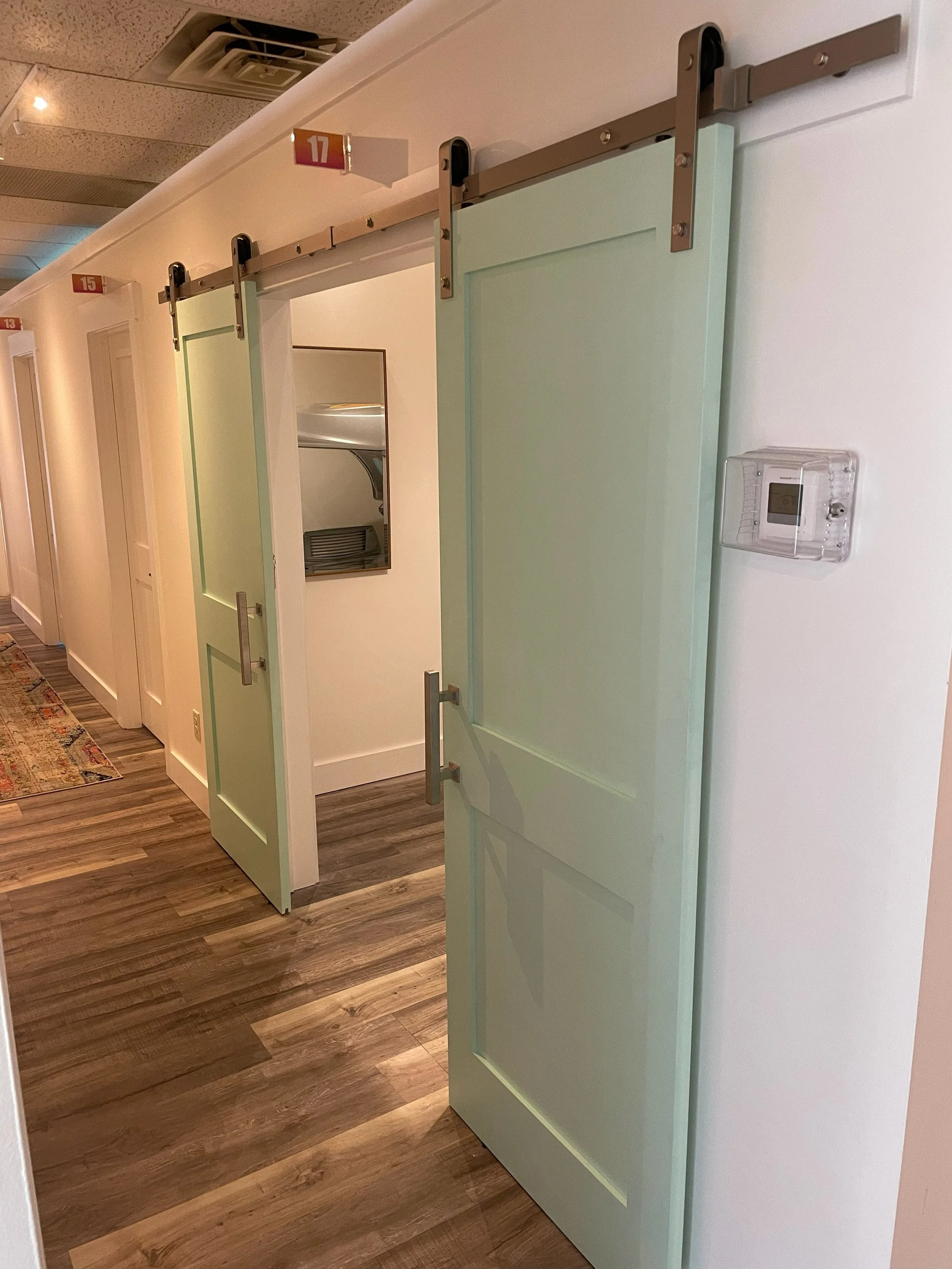 A hallway with avocado green sliding barn doors on a track system. Each door has a metallic handle. The hallway has numbered signs above each doorway, with numbers 13, 15, and 17 visible. There is wood-patterned flooring and part of a colorful rug on