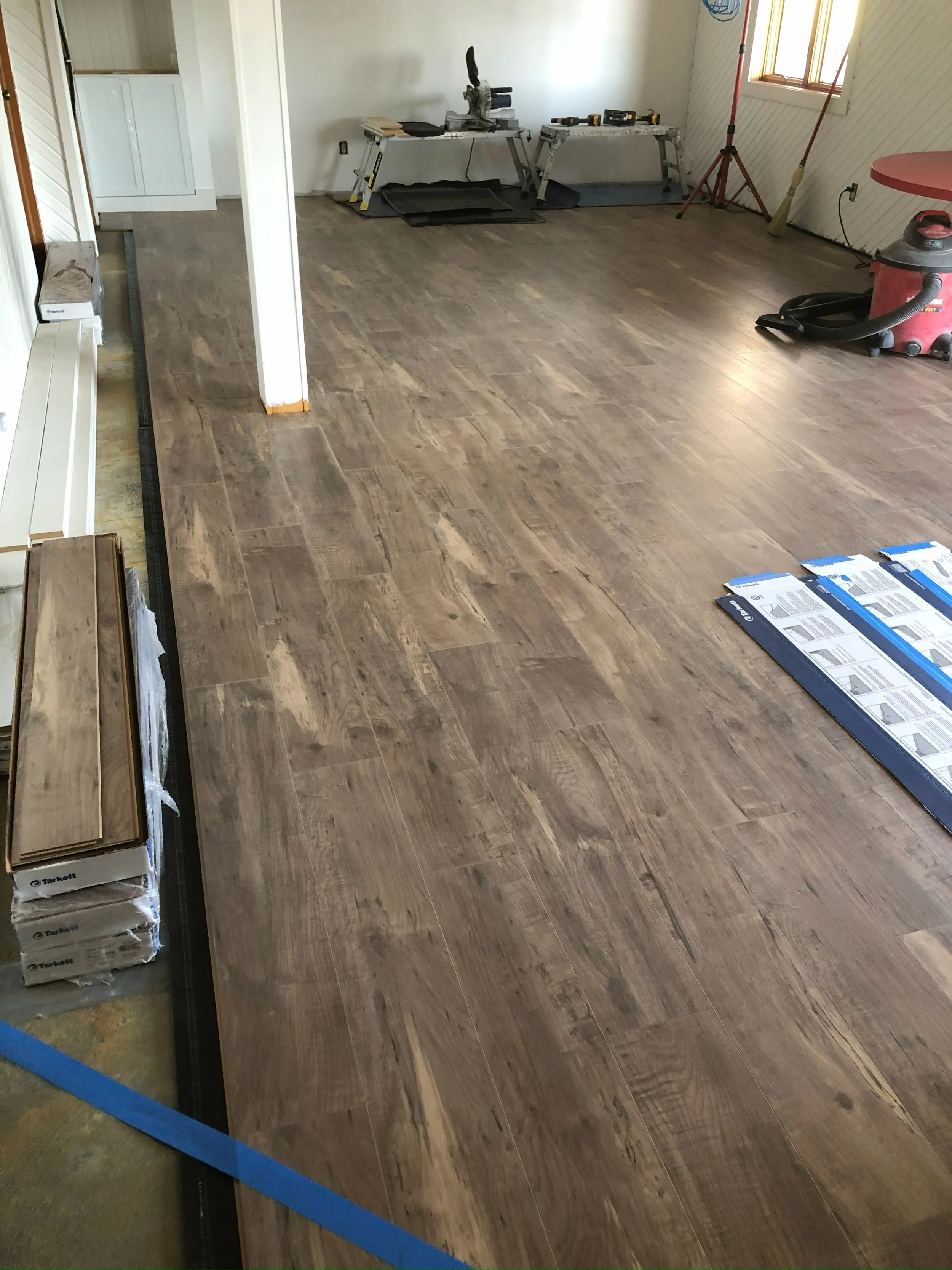 Living room with newly installed wooden flooring, construction tools, and supplies, including a miter saw on a table, a shop vacuum, and boxes of flooring materials.