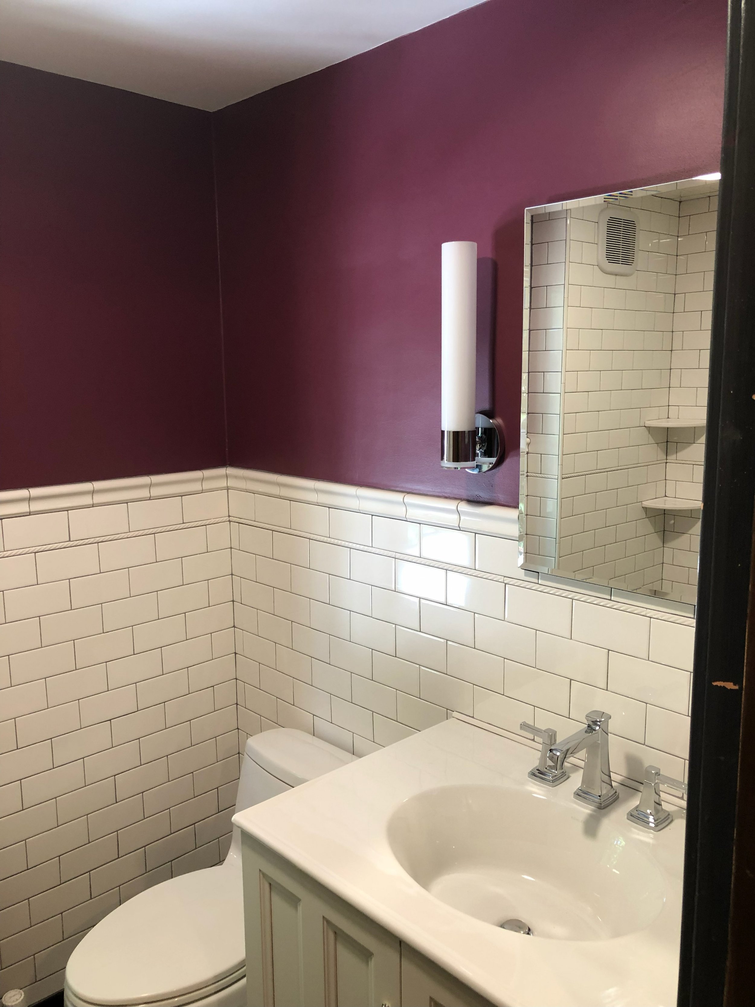 Small bathroom with purple painted upper wall, white subway tile lower wall, featuring a white toilet, a white vanity with a sink, a silver faucet, a rectangular mirror, and a vertical white wall-mounted light fixture.