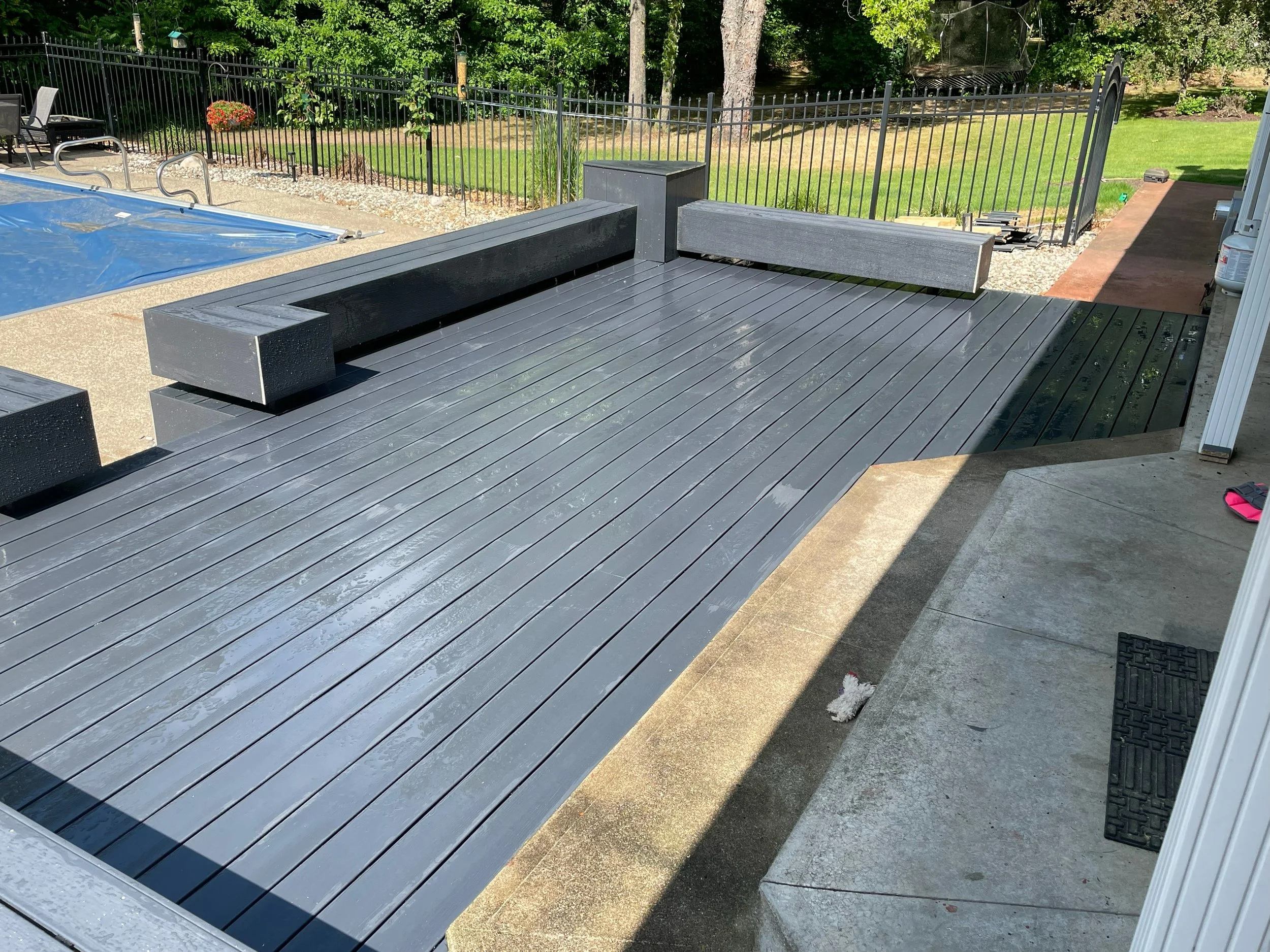 A newly built backyard deck with black painted wooden planks, surrounded by a black metal fence, with a pool covered with a blue cover on one side and green trees and lawn in the background.