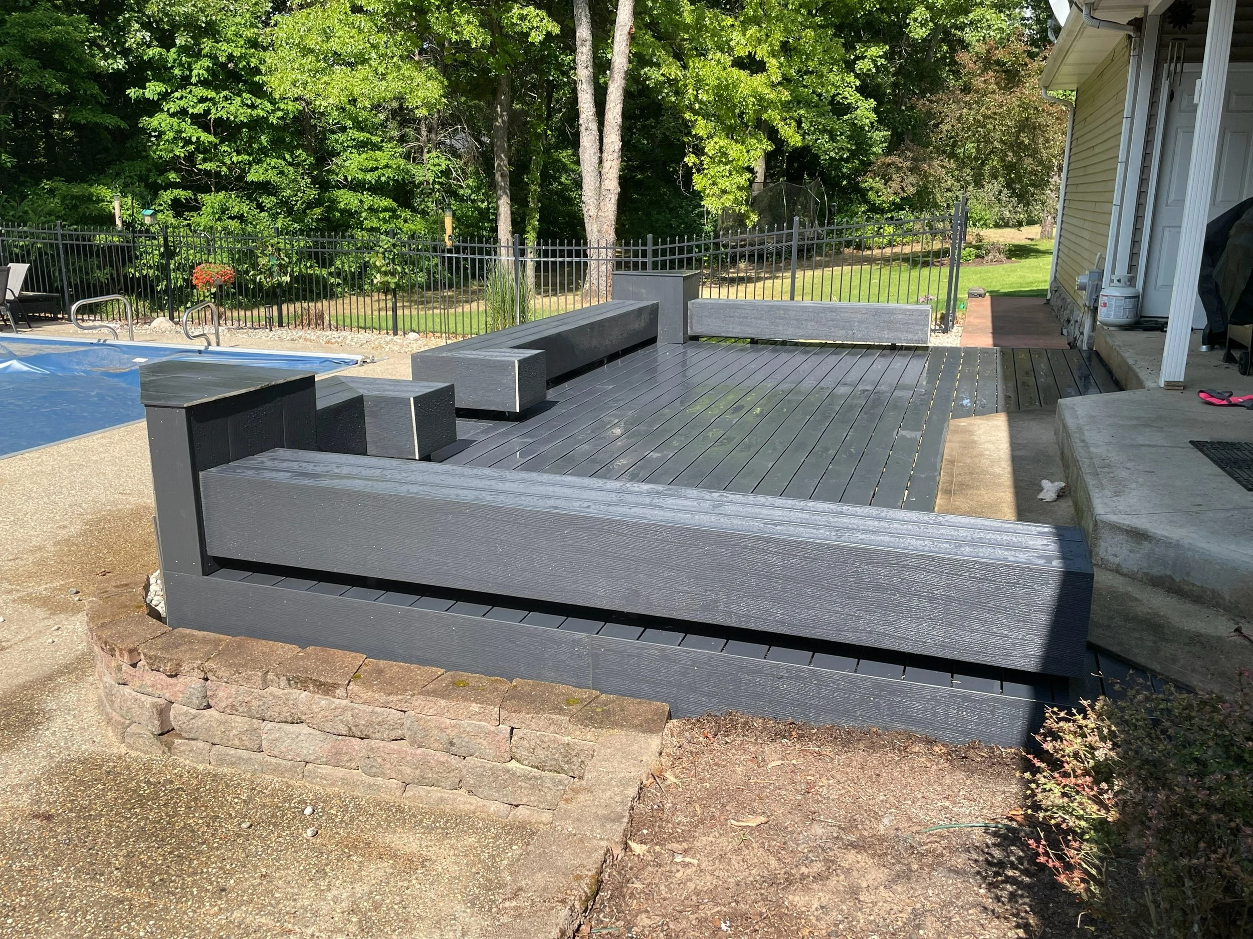 Backyard patio with black wooden furniture, including a low table and built-in benches, surrounded by a brick border. There is a pool with a cover on the left and a fence with trees in the background. A house with beige siding is on the right side.