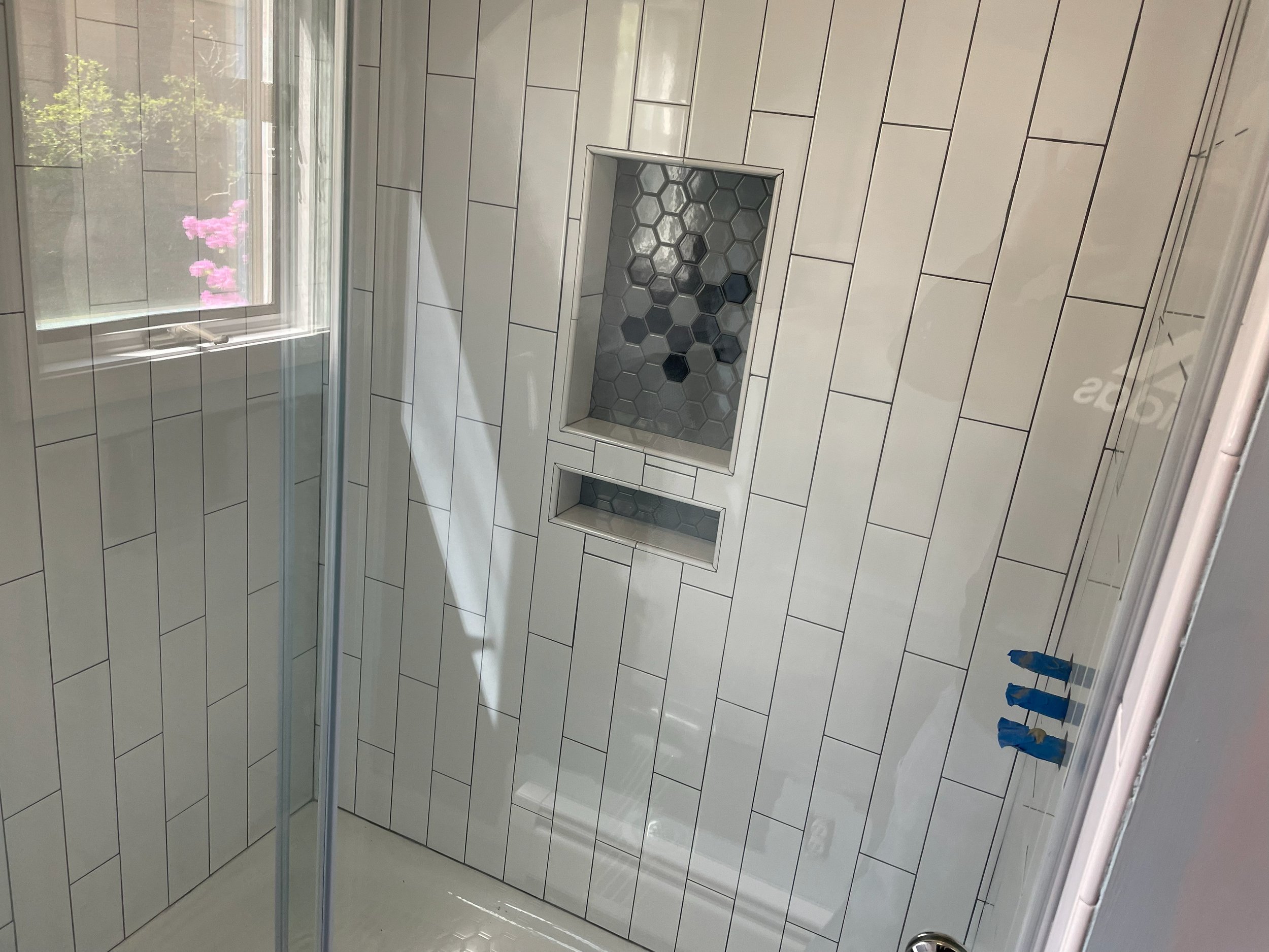 A modern shower with white subway tiles and a built-in niche with hexagon tiles, next to a window with pink flowers outside.