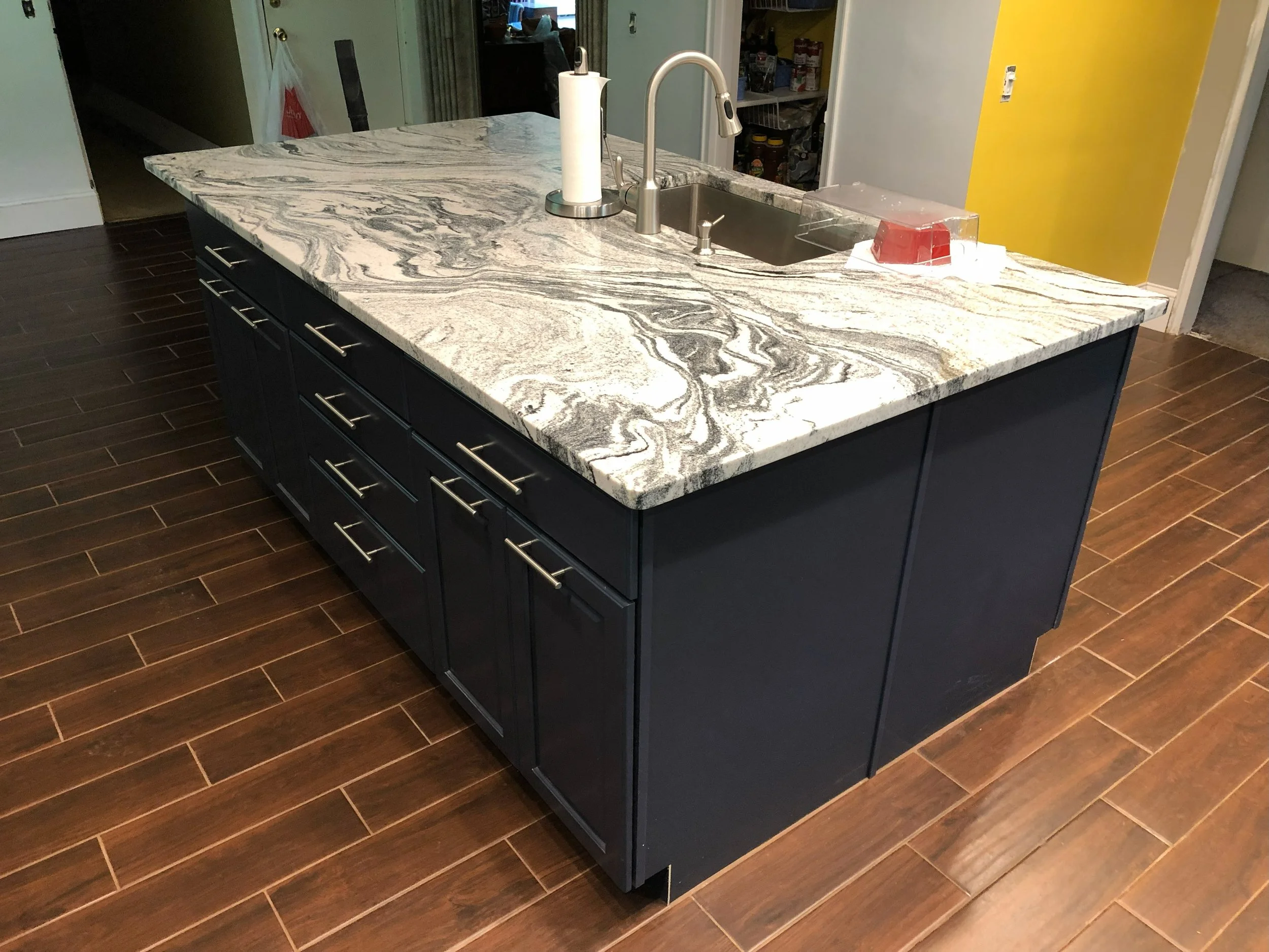 Kitchen island with a marble countertop, cabinetry with silver handles, a sink with a faucet, paper towel roll, and a plastic container with red items. Hardwood floor and a partially yellow painted wall in the background.