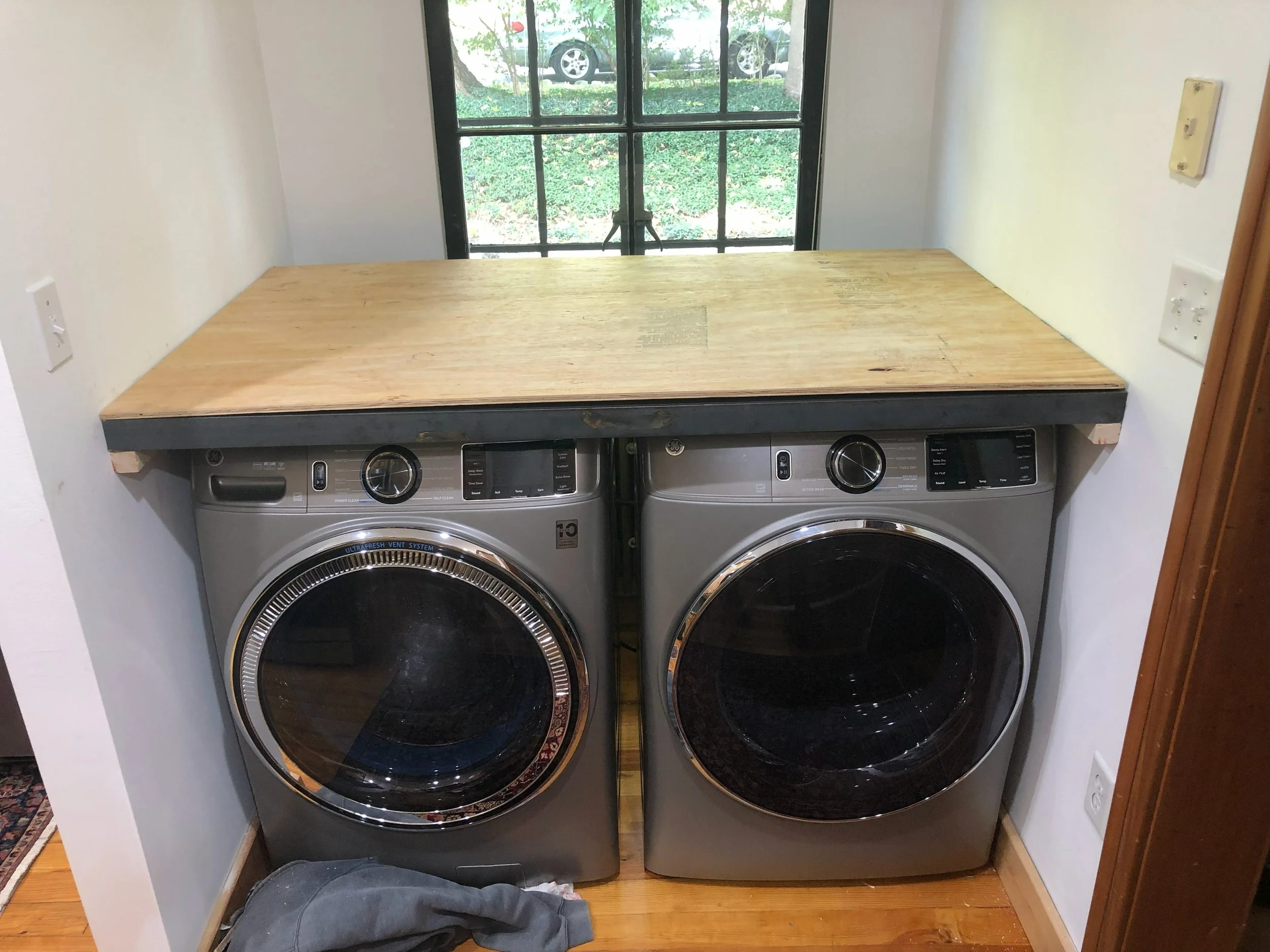 Front-loading washing machine and dryer side by side under a wooden countertop in a laundry room, with a window above.