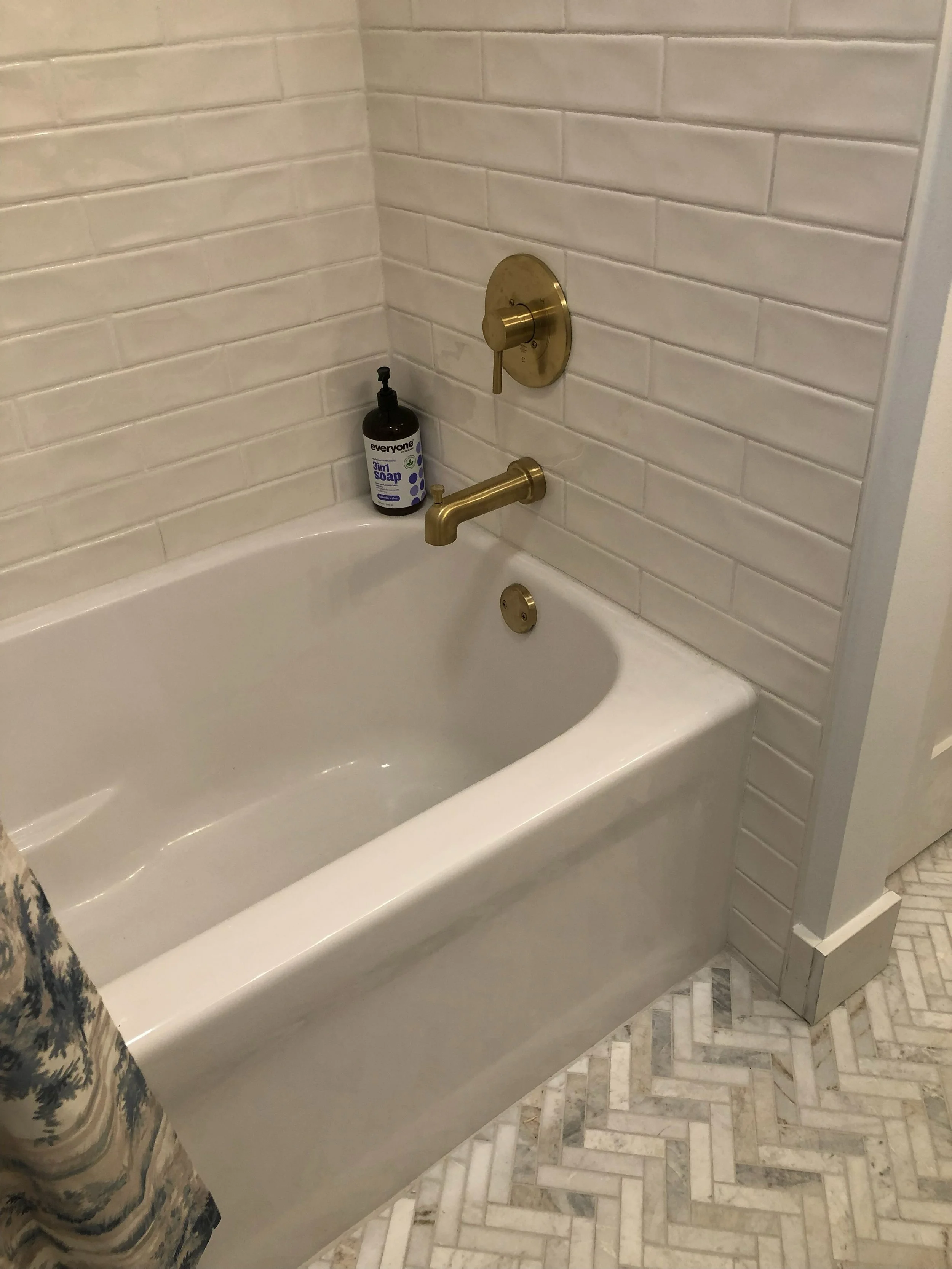 Bathroom bathtub with gold faucet and shower controls, soap dispenser on the edge, and patterned tile flooring.