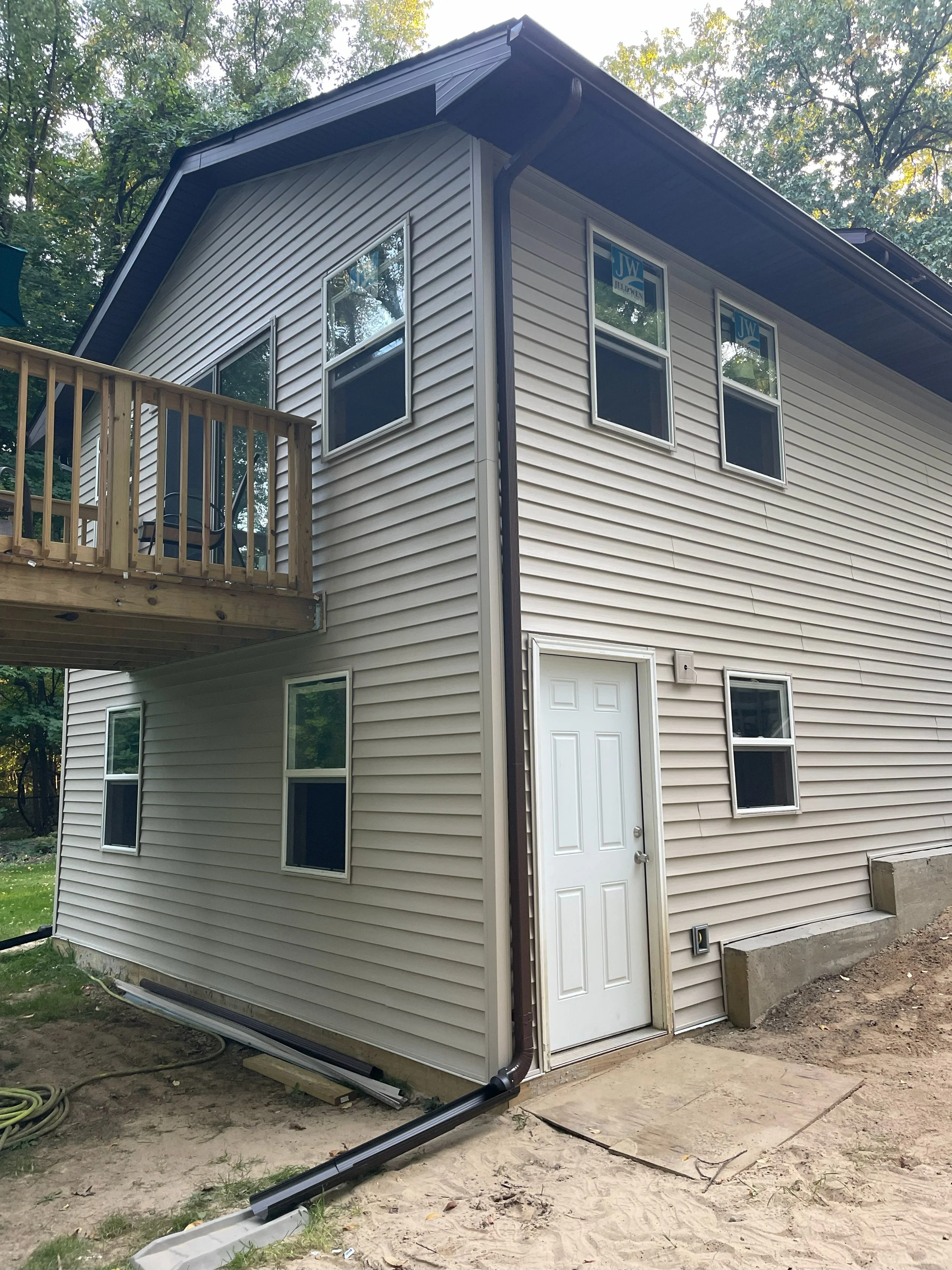 A two-story house with beige vinyl siding, multiple windows, a white door, and a small wooden balcony. The house has a dark brown roof and is situated in a wooded area.