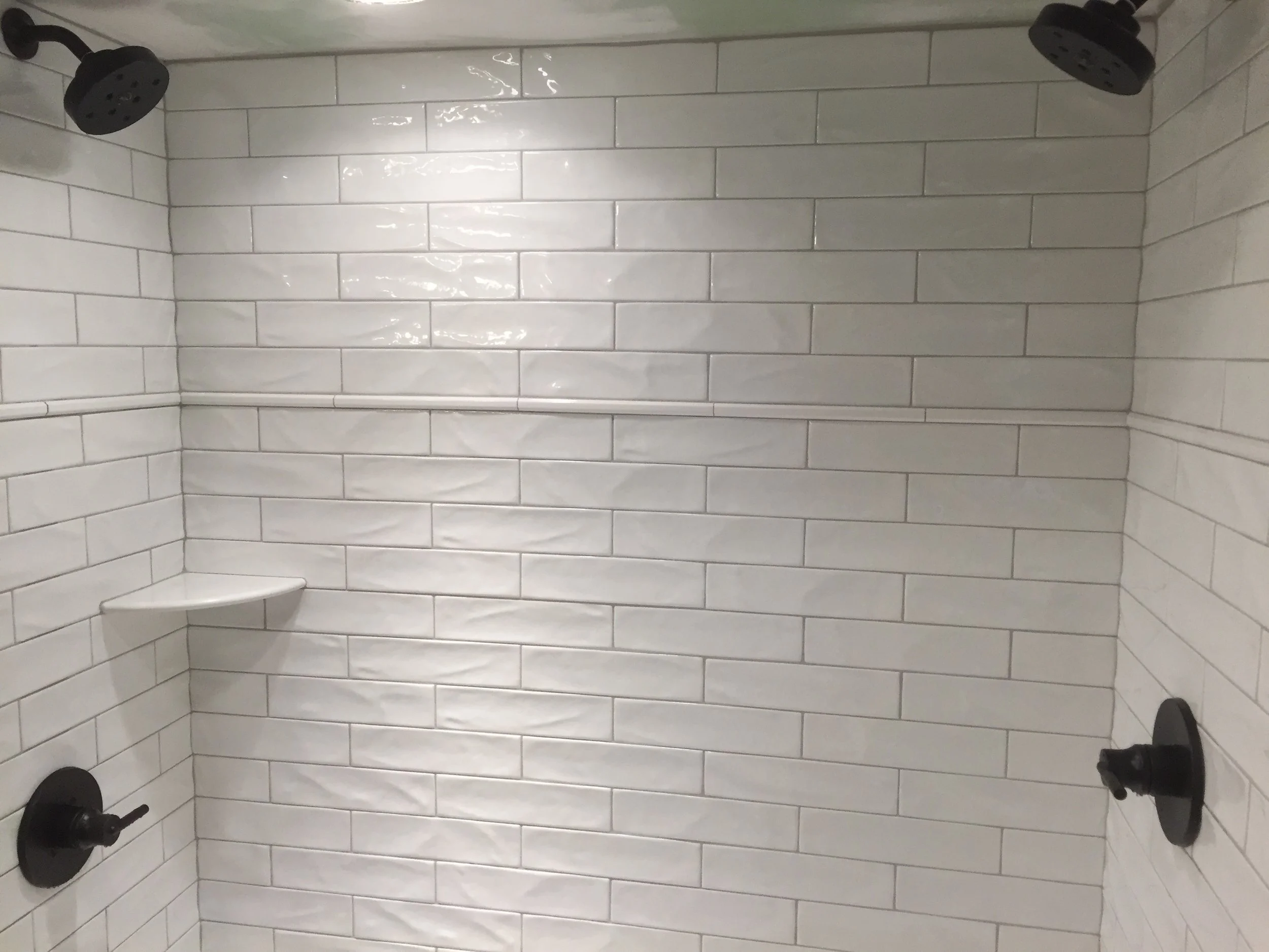 A walk-in shower with white subway tile walls, two black showerheads on the ceiling, a small corner shelf, and black fixtures.