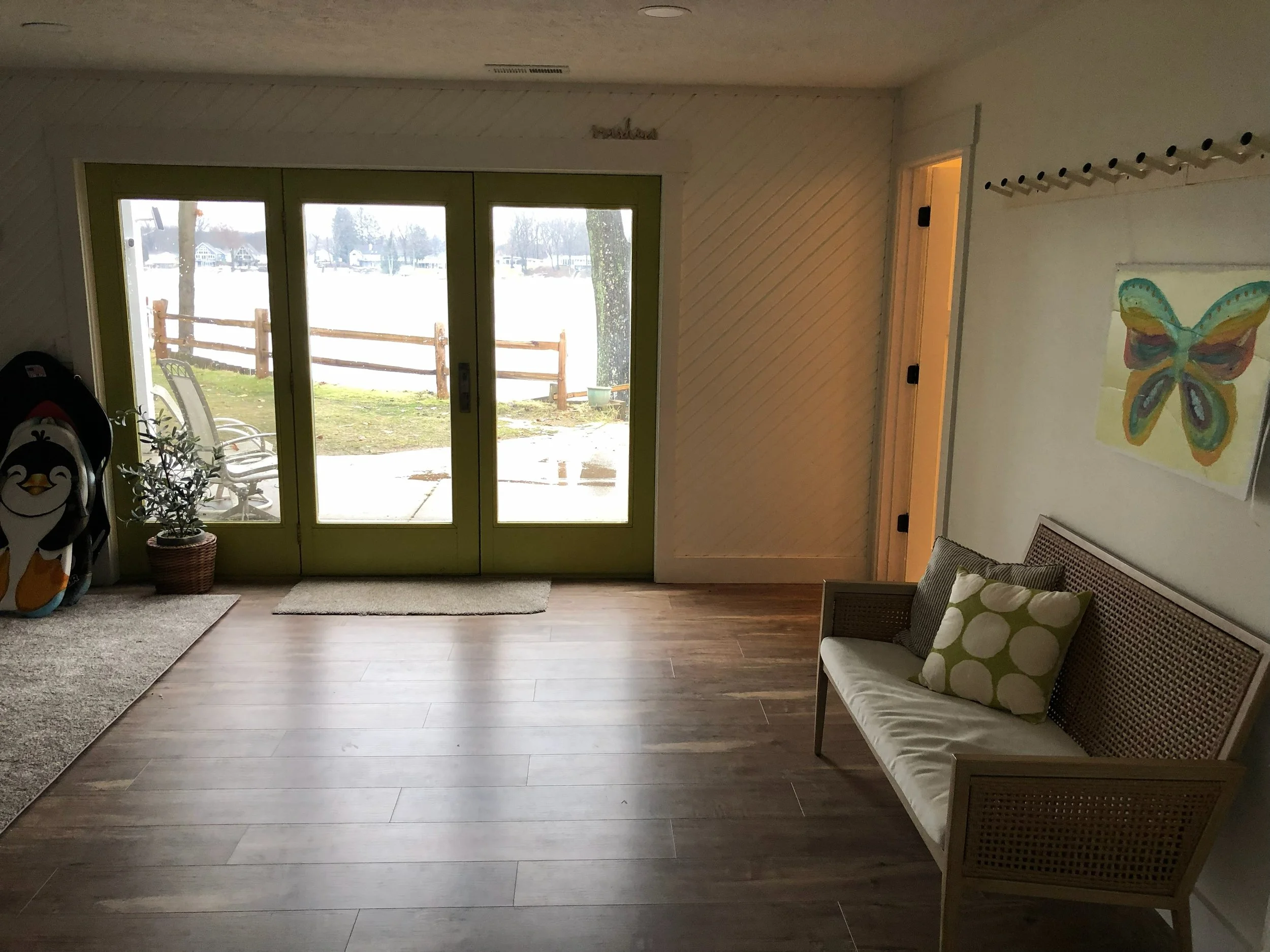 Living room with large glass doors leading outside, a bench with pillows, a colorful butterfly painting on the wall, and a few items near the door.