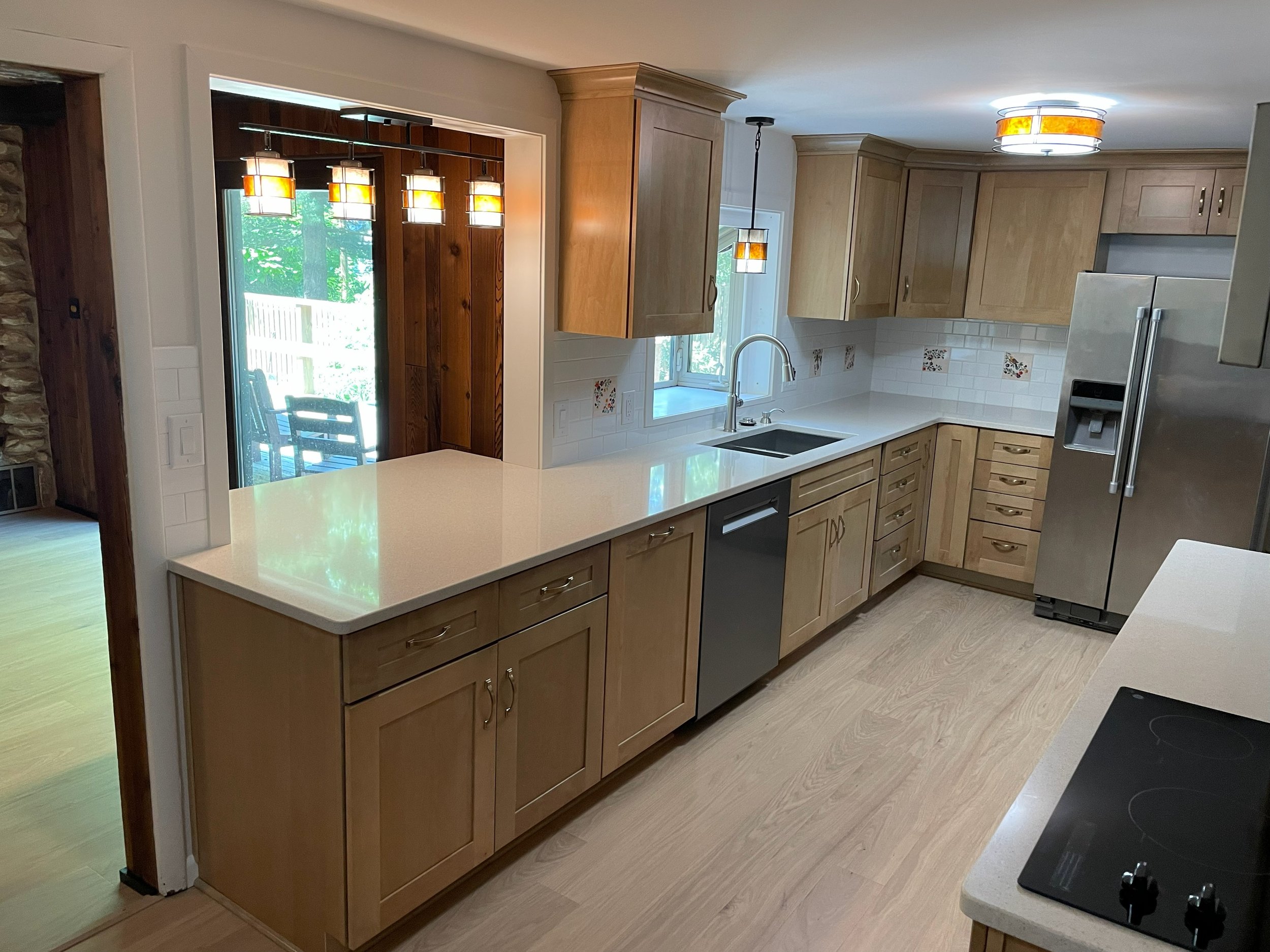 Kitchen with light wood cabinets, white countertops, stainless steel refrigerator, black dishwasher, and an electric stove. There is a window above the sink and a glass sliding door leading to an outdoor dining area. Pendant lighting is over the coun