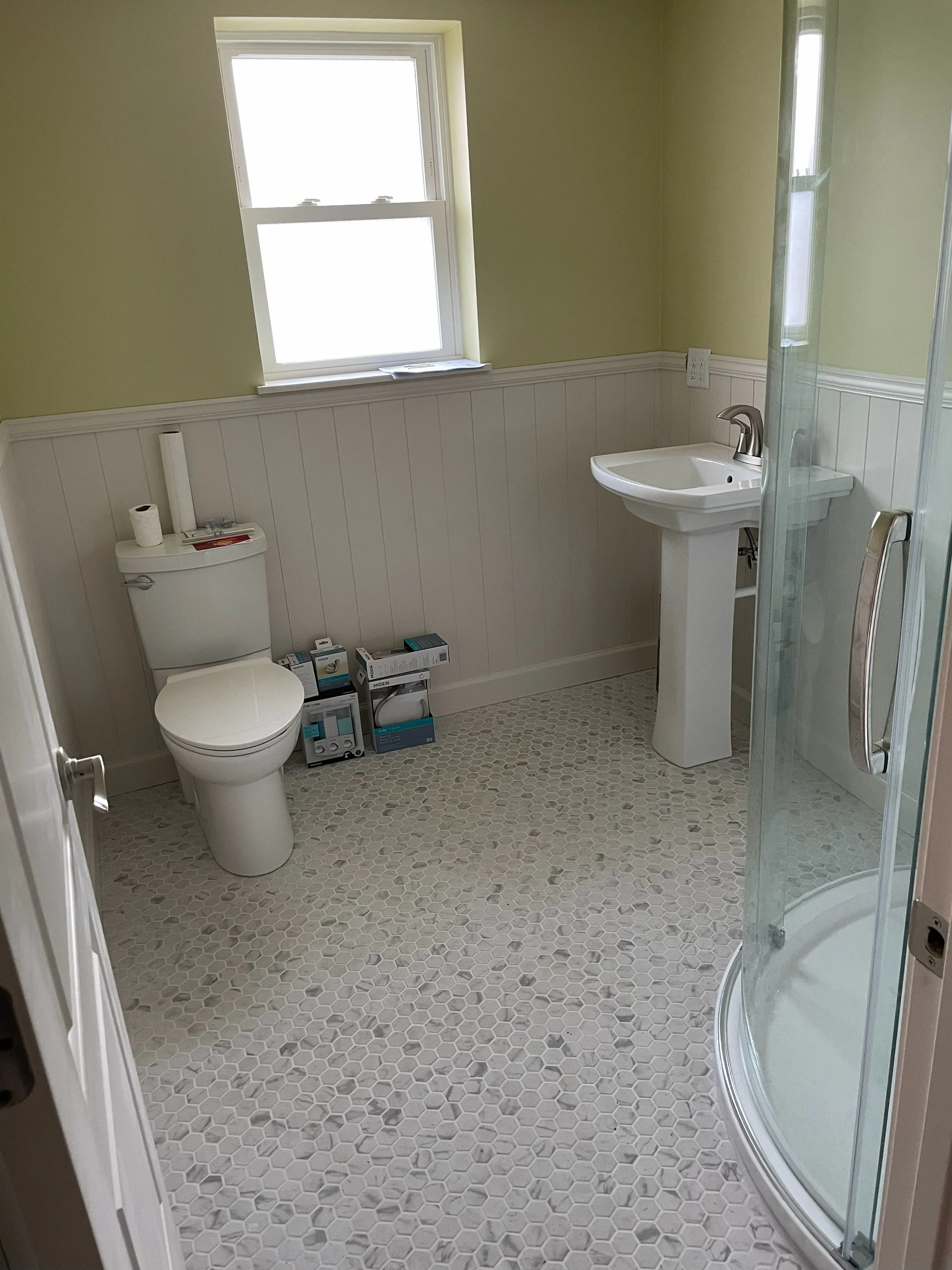 A bathroom with a toilet, a small sink, a corner shower, and a window. The floor is tiled with small hexagonal tiles, and the walls have a white beadboard wainscot with light green paint above.