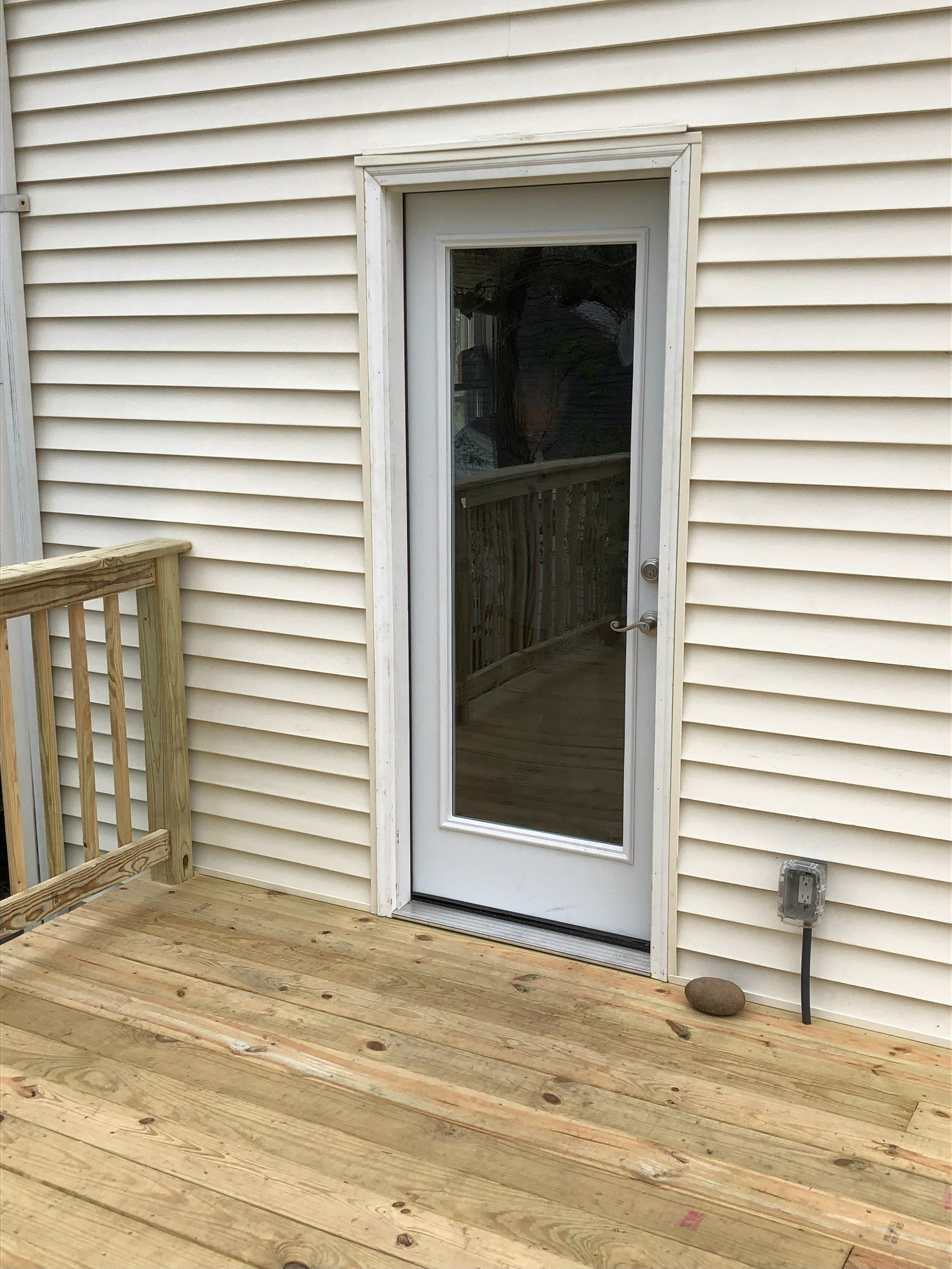 A wooden deck with a railing, beige vinyl siding wall, a white door with a glass panel, and an outdoor electrical outlet with a rock placed in front of it.